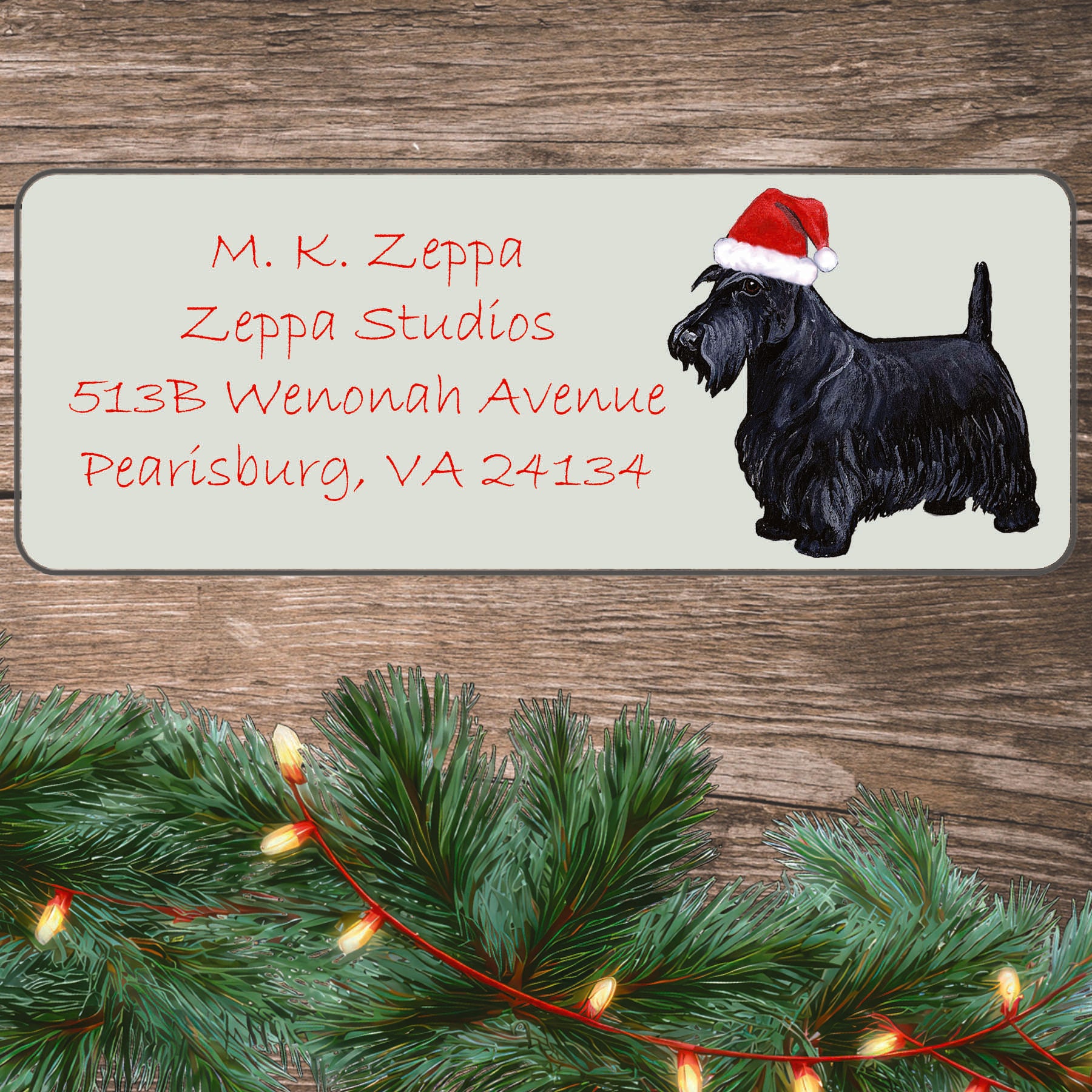 An illustration of a Scottish Terrier wearing a Santa Hat on an ivory return address label set off by a wood surface with holiday greenery background.