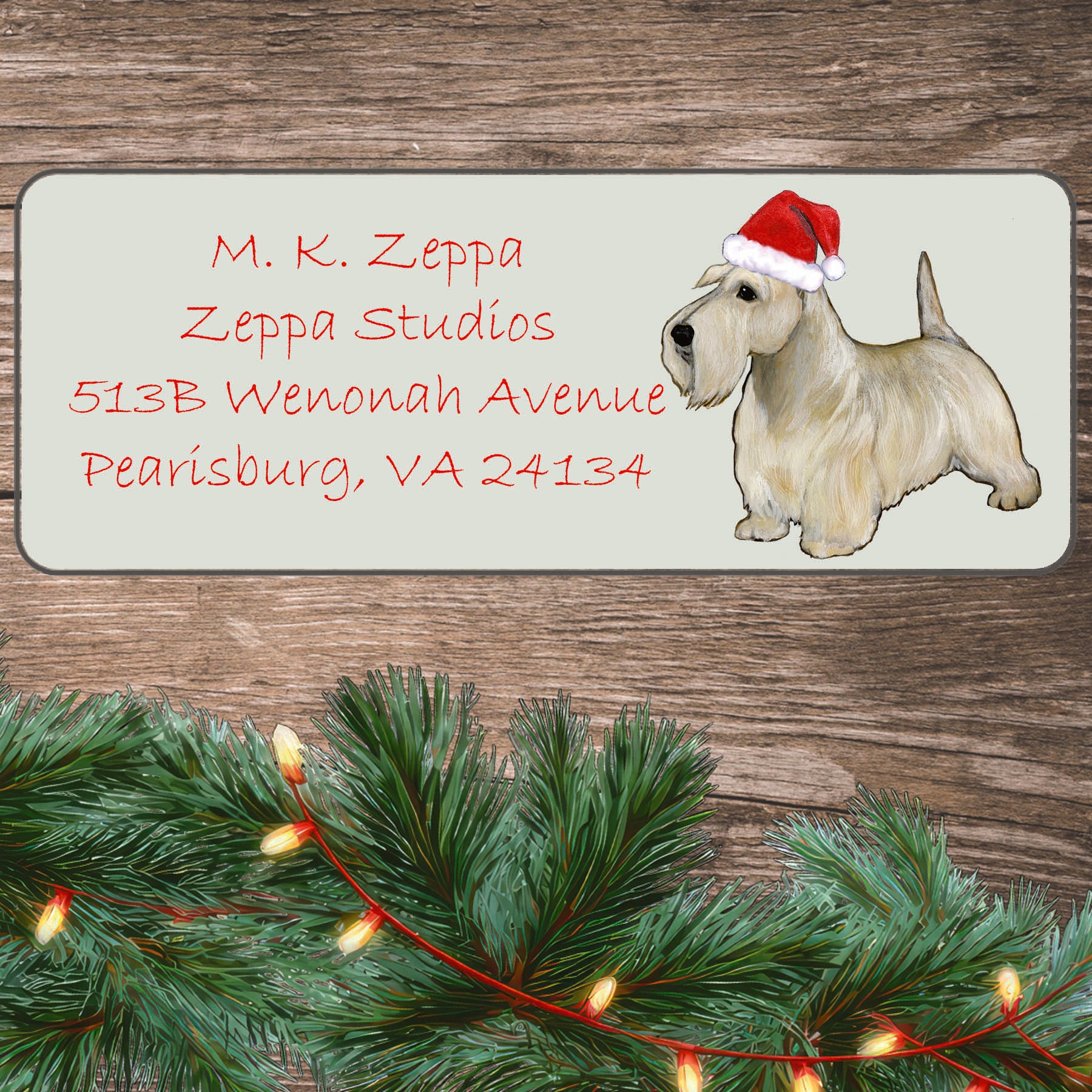 An illustration of a wheaten Scottish Terrier wearing a Santa Hat on an ivory return address label set off by a wood surface with holiday greenery background.