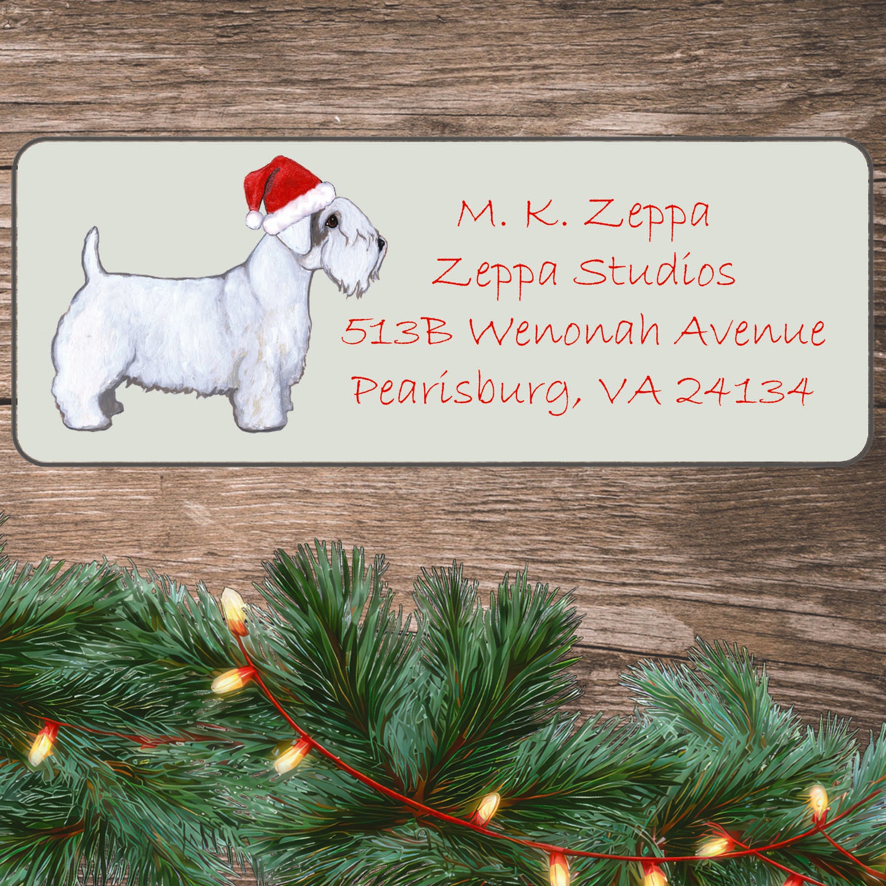 An illustration of a Sealyham Terrier wearing a Santa Hat on an ivory return address label set off by a wood surface with holiday greenery background.