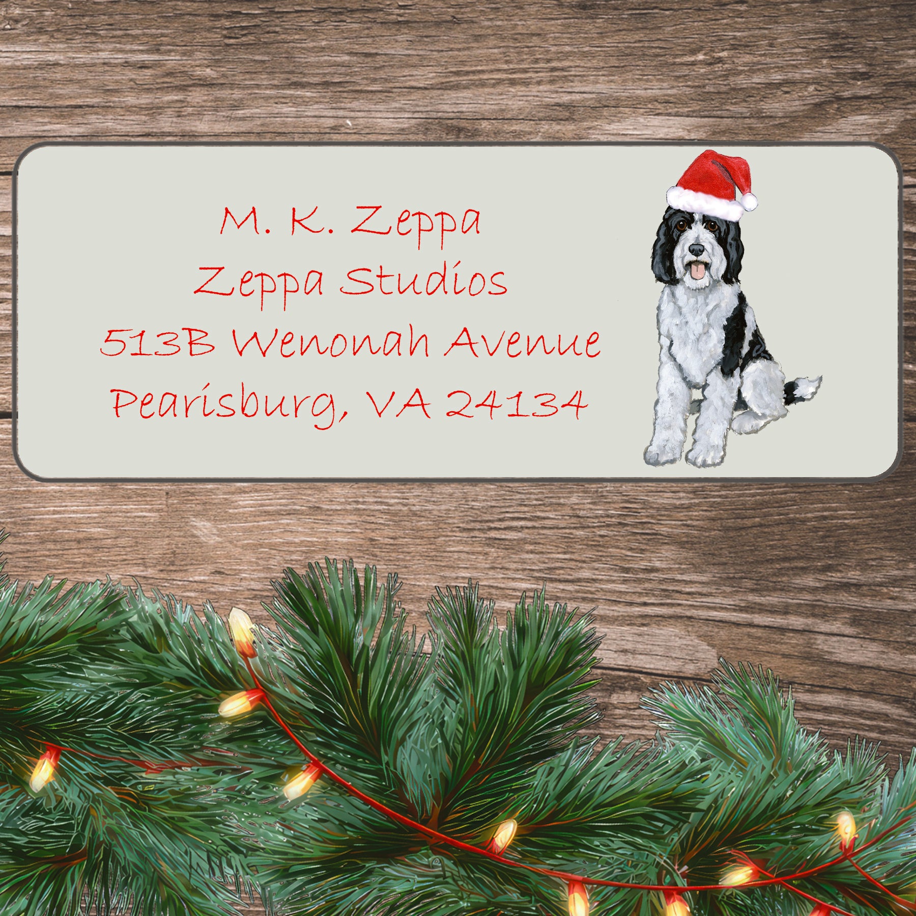 A Sheepadoodle in a Santa Hat illustration on an ivory return address label set off by a wood surface with holiday greenery background.