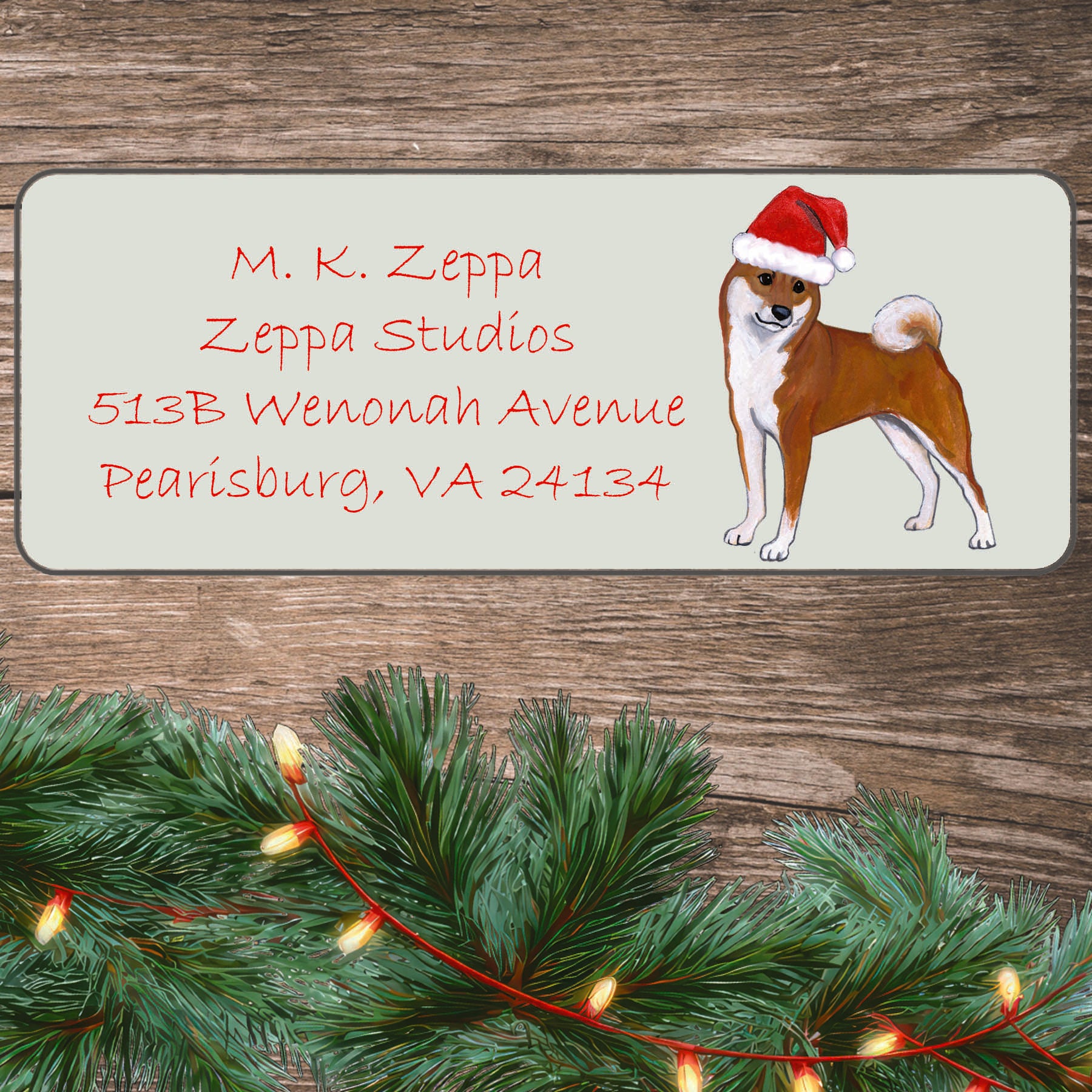 An illustration of a Shiba Inu wearing a Santa Hat on an ivory return address label set off by a wood surface with holiday greenery background.