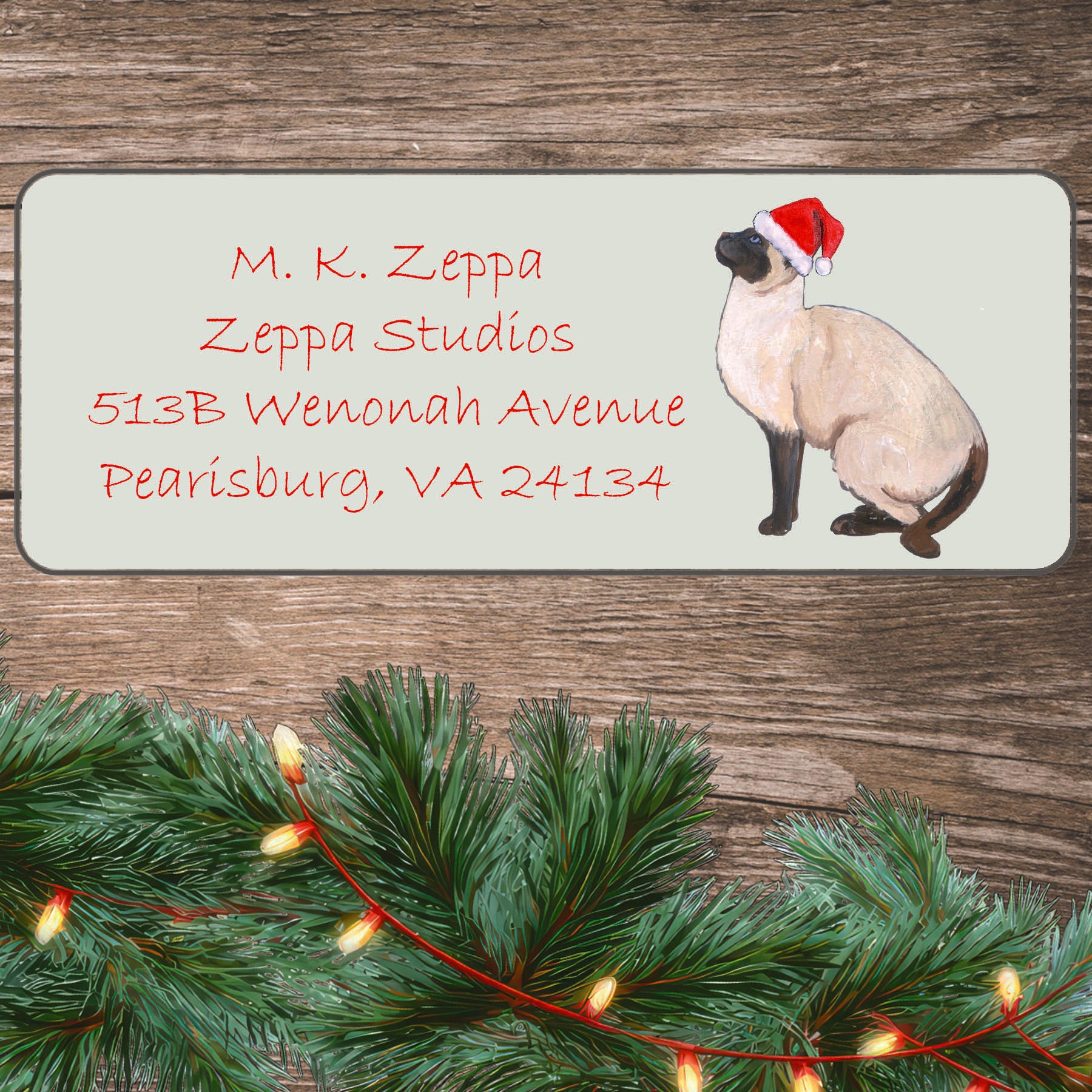 An illustration of a Siamese Cat wearing a Santa Hat on an ivory return address label set off by a wood surface with holiday greenery background.