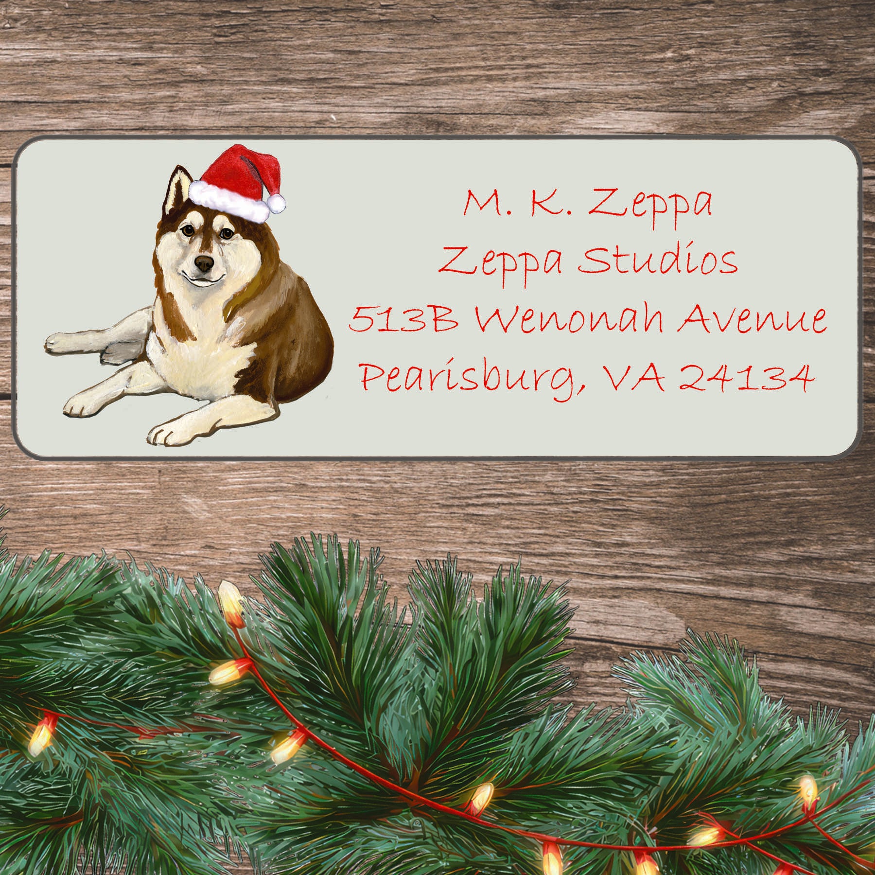 An illustration of a brown and white Siberian Husky wearing a Santa Hat on an ivory return address label set off by a wood surface with holiday greenery background.