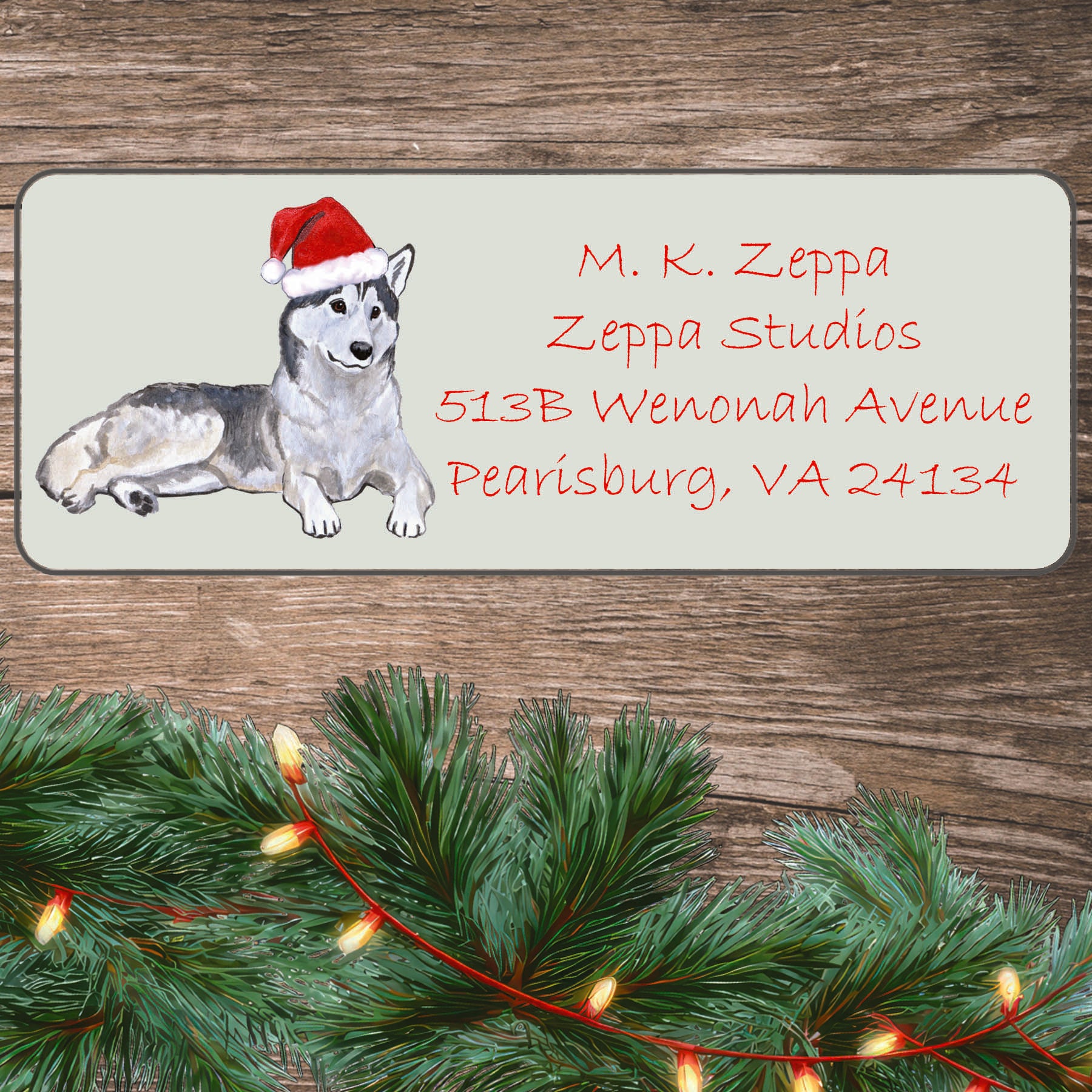 An illustration of a Siberian Husky wearing a Santa Hat on an ivory return address label set off by a wood surface with holiday greenery background.