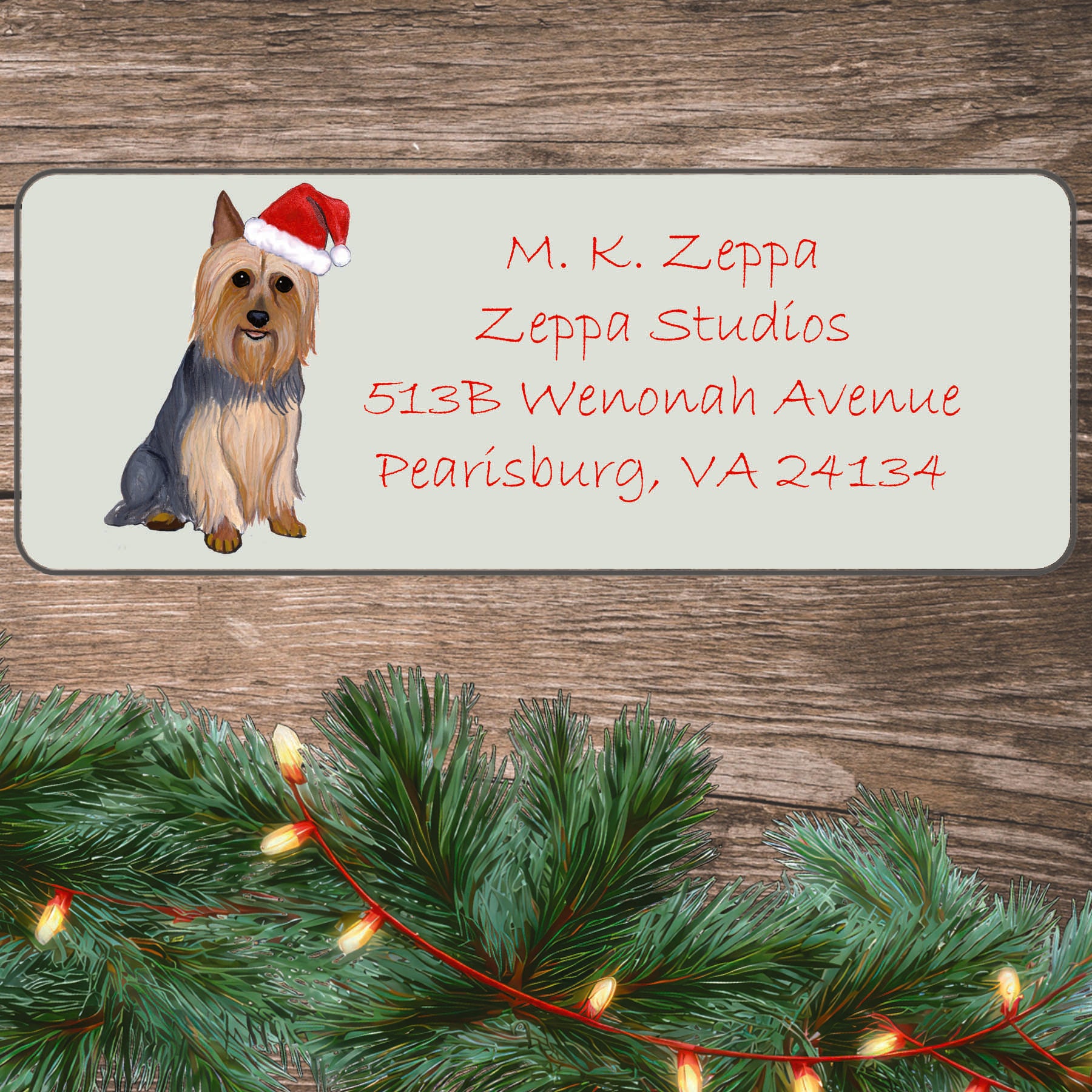 An illustration of a Silky Terrier wearing a Santa Hat on an ivory return address label set off by a wood surface with holiday greenery background.
