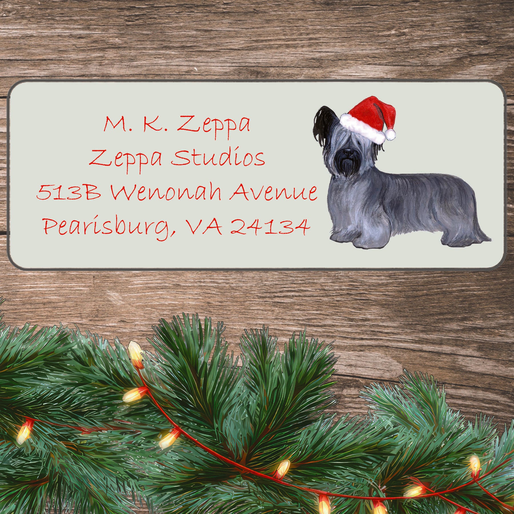 An illustration of a Skye Terrier wearing a Santa Hat on an ivory return address label set off by a wood surface with holiday greenery background.