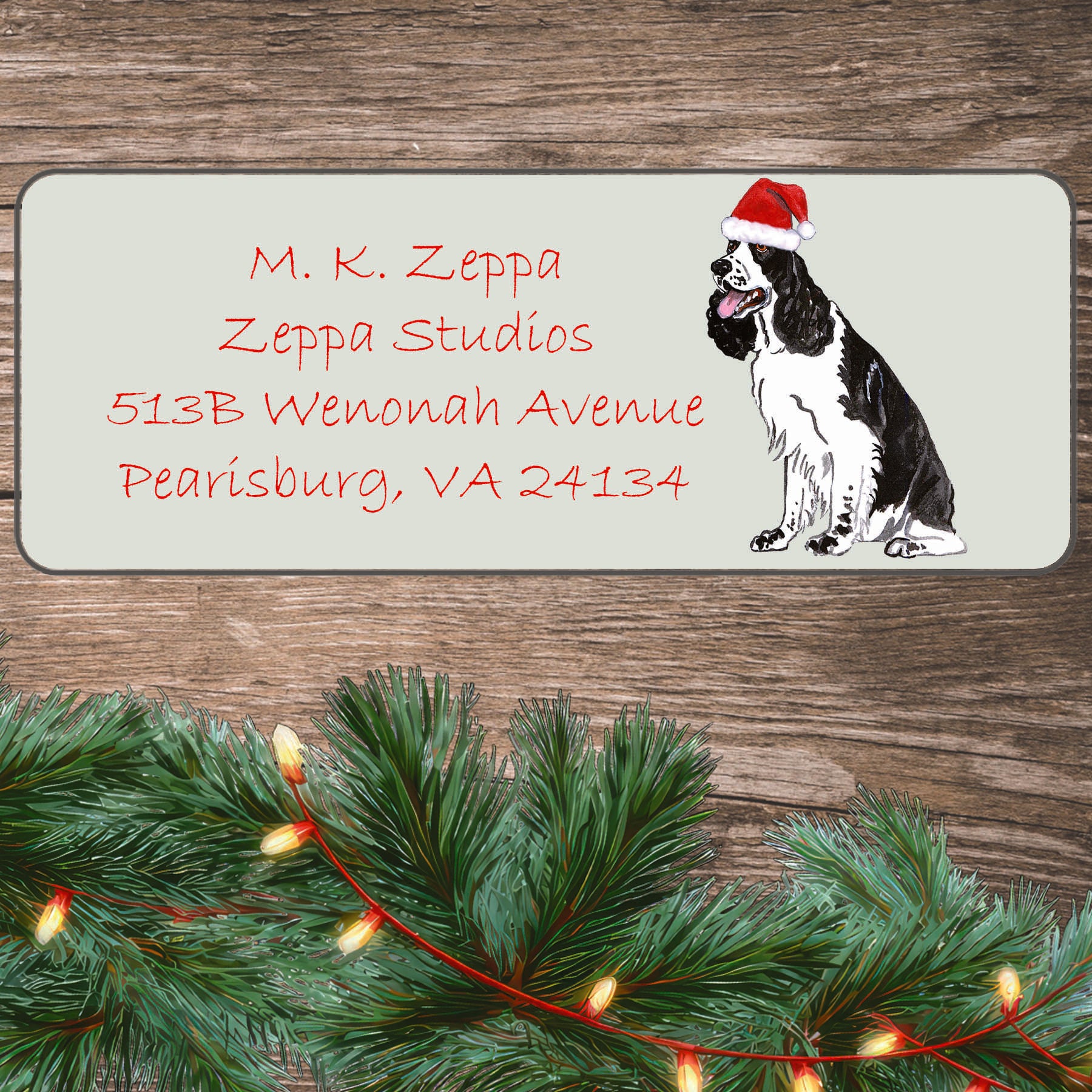 An illustration of a black and white Springer Spaniel wearing a Santa Hat on an ivory return address label set off by a wood surface with holiday greenery background.