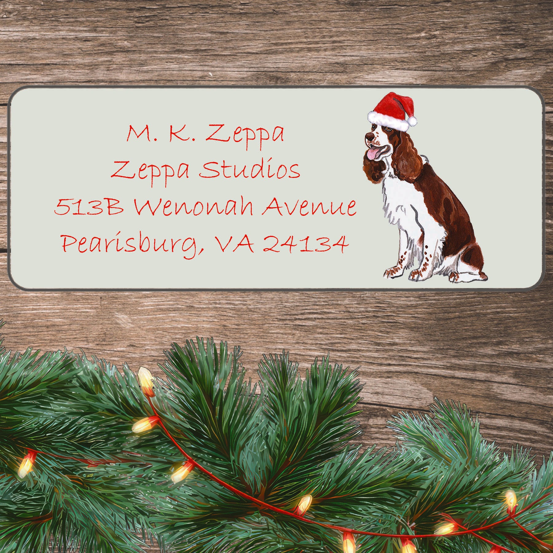 An illustration of a Springer Spaniel wearing a Santa Hat on an ivory return address label set off by a wood surface with holiday greenery background.