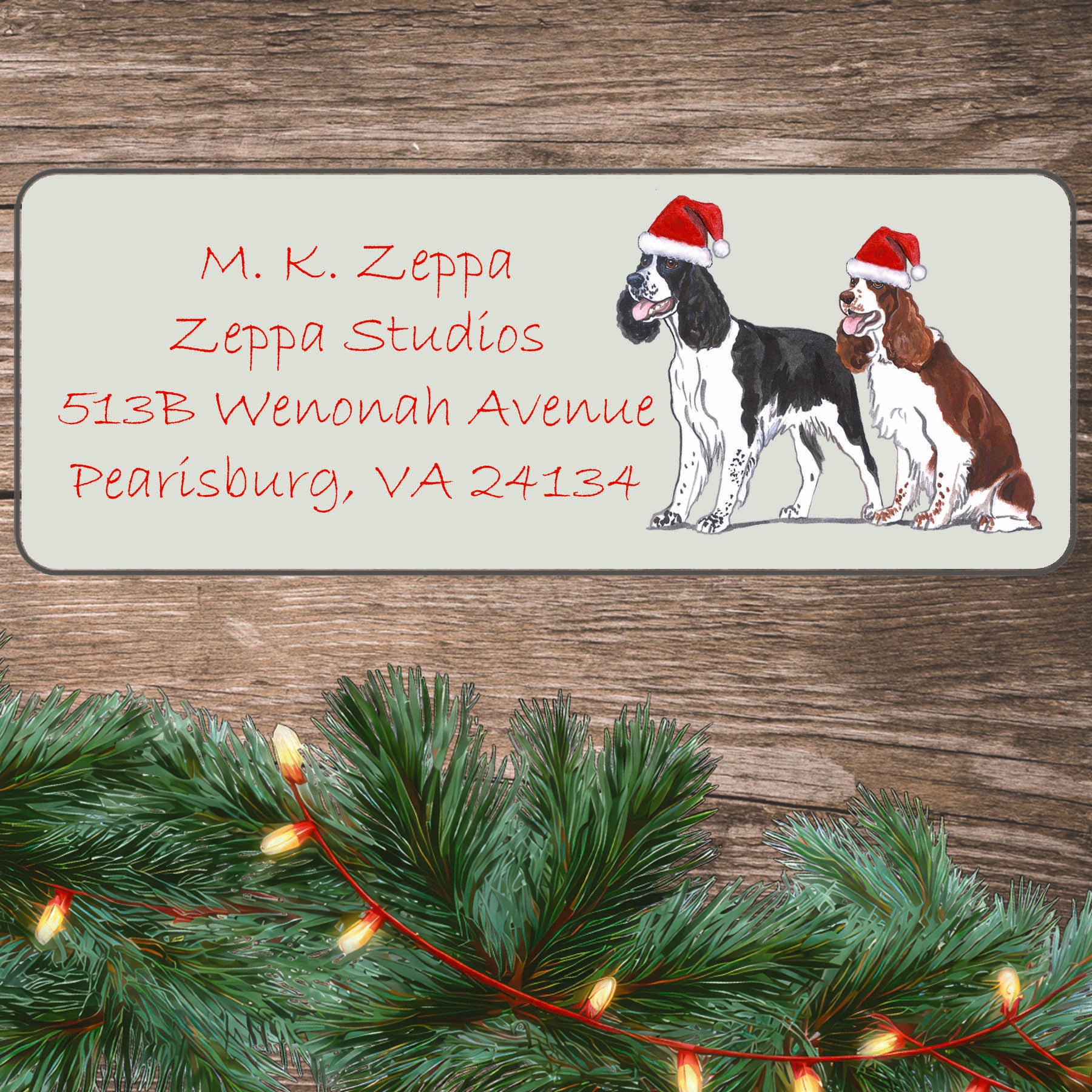 An illustration of a pair of Springer Spaniels wearing Santa Hats on an ivory return address label set off by a wood surface with holiday greenery background.
