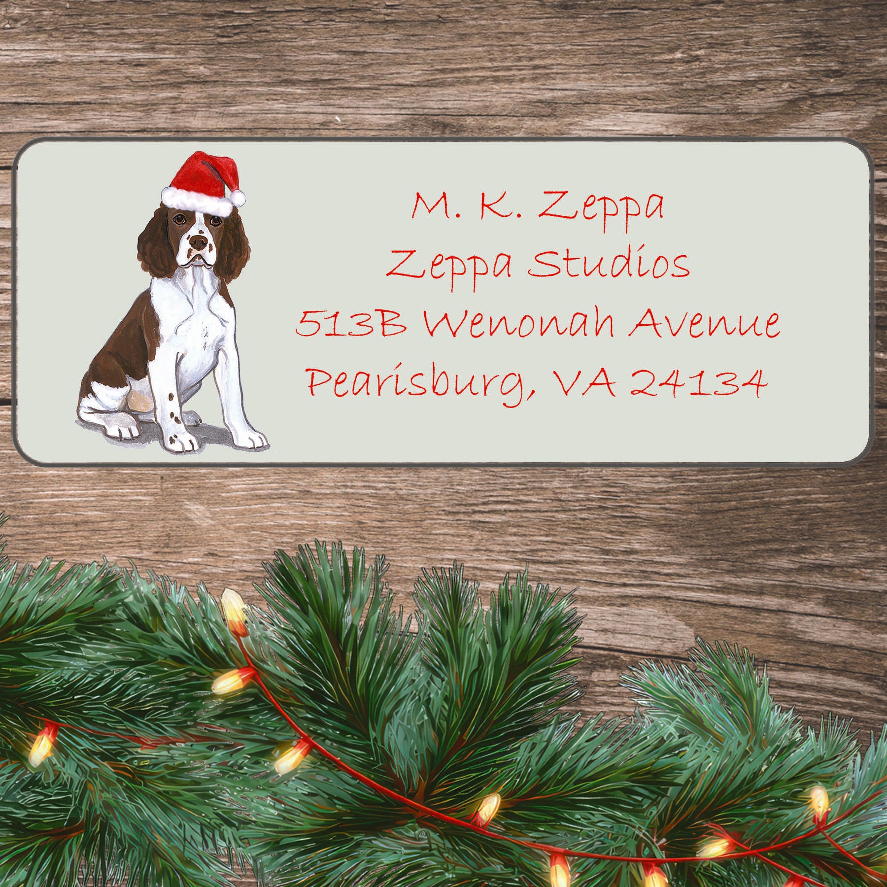 An illustration of a Springer Spaniel Puppy wearing a Santa Hat on an ivory return address label set off by a wood surface with holiday greenery background.