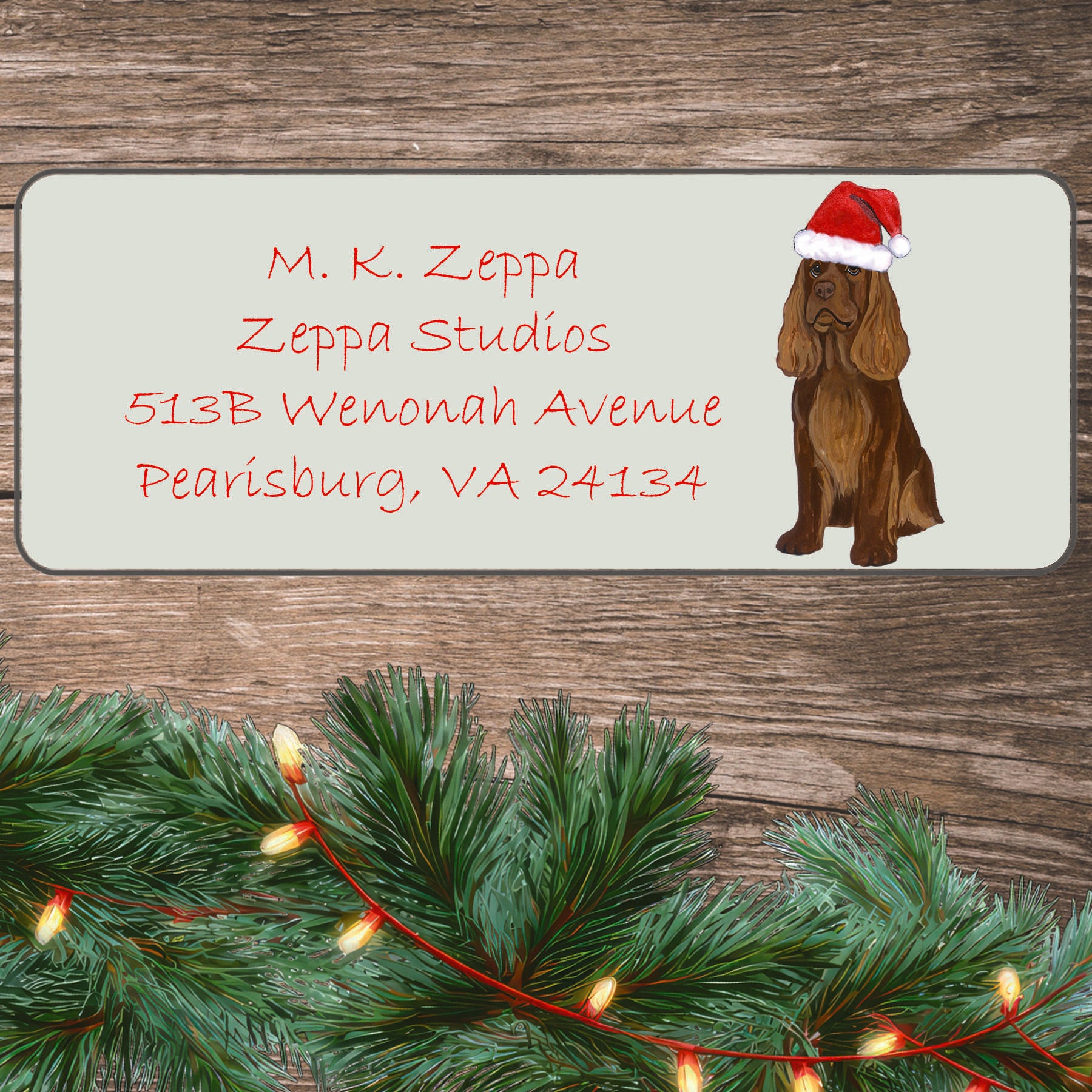 An illustration of a Sussex Spaniel wearing a Santa Hat on an ivory return address label set off by a wood surface with holiday greenery background.
