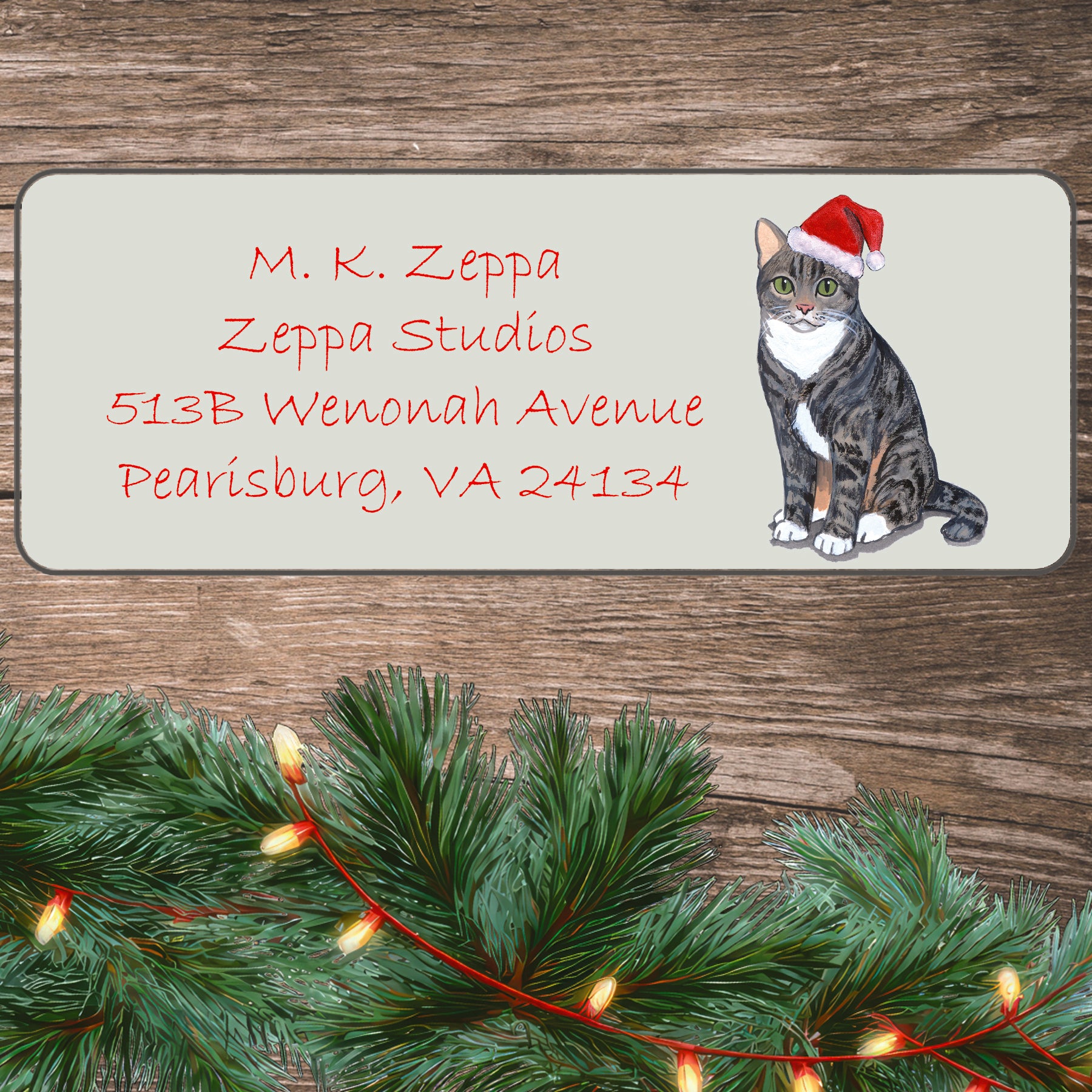 An illustration of a  grey Tabby Cat wearing a Santa Hat on an ivory return address label set off by a wood surface with holiday greenery background.