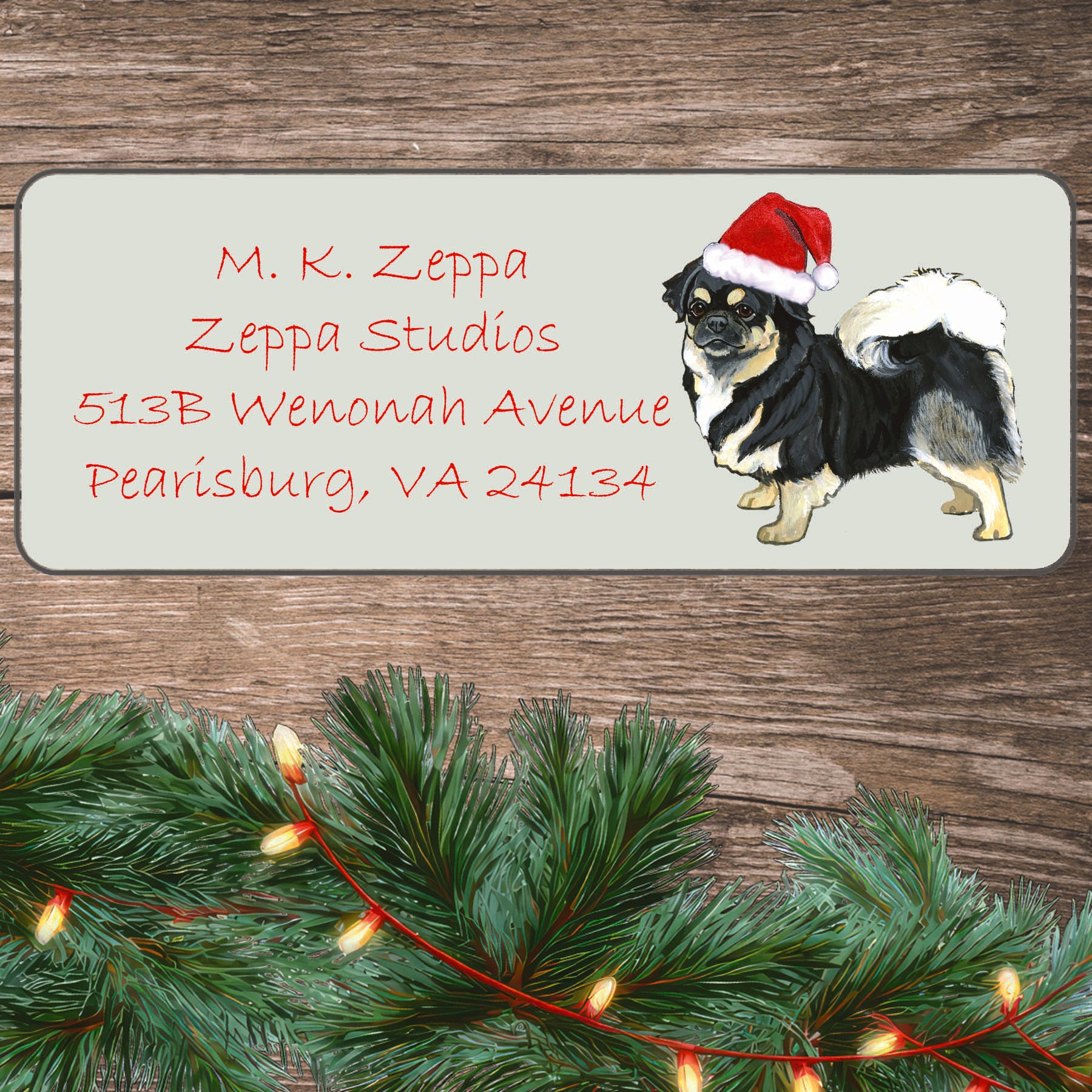 An illustration of a black and tan Tibetan Spaniel wearing a Santa Hat on an ivory return address label set off by a wood surface with holiday greenery background.