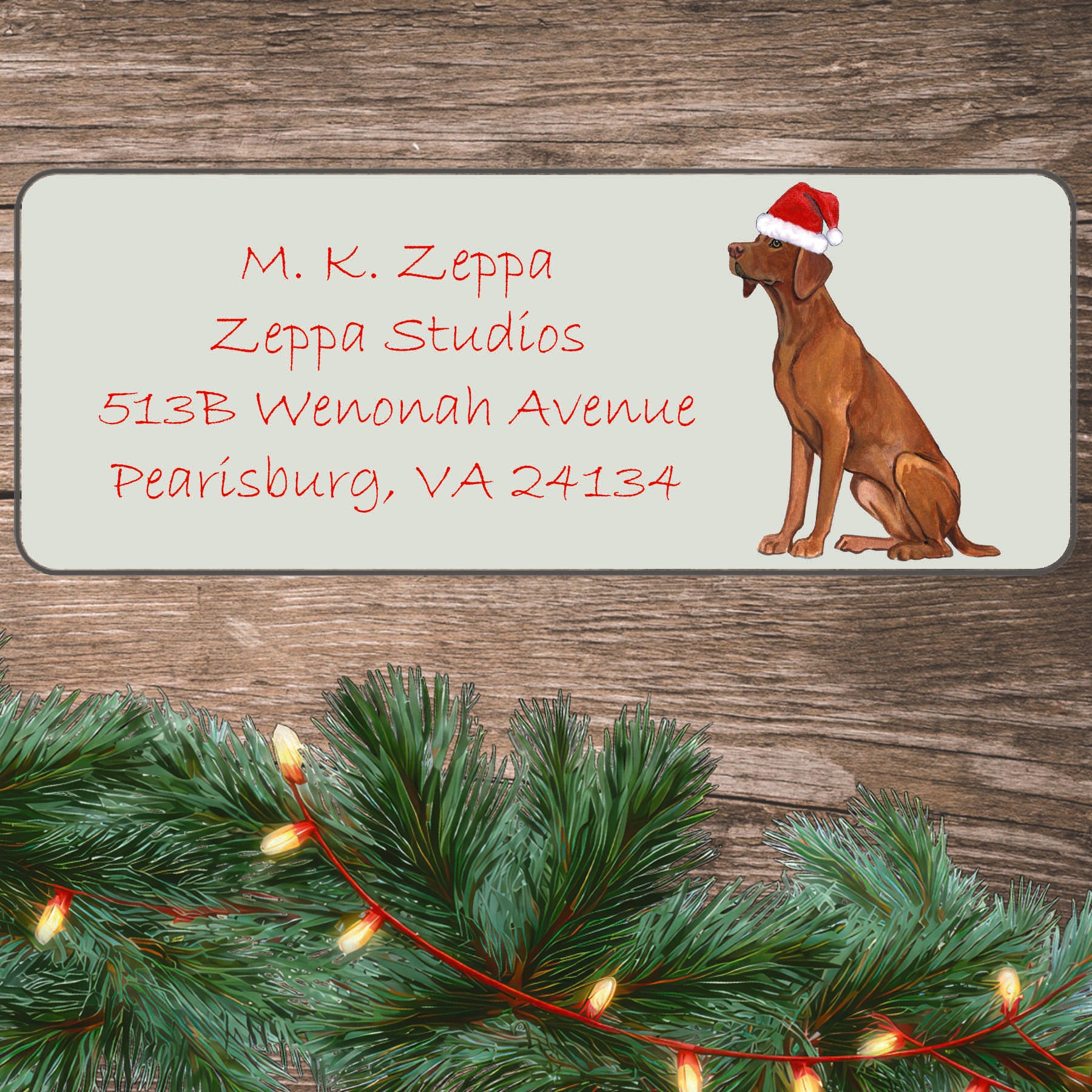 An illustration of a Vizsla wearing a Santa Hat on an ivory return address label set off by a wood surface with holiday greenery background.