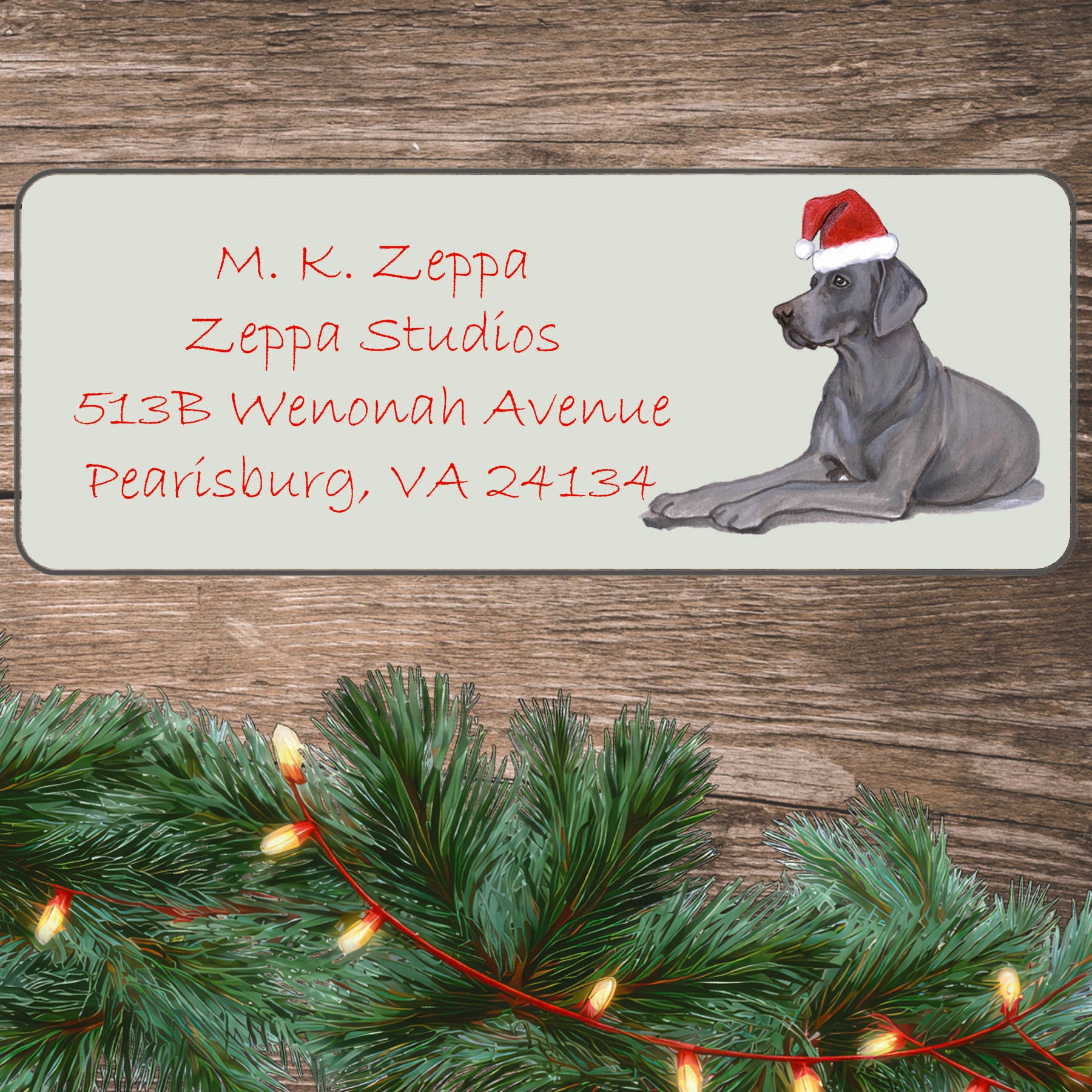 An illustration of a Weimaraner wearing a Santa Hat on an ivory return address label set off by a wood surface with holiday greenery background.