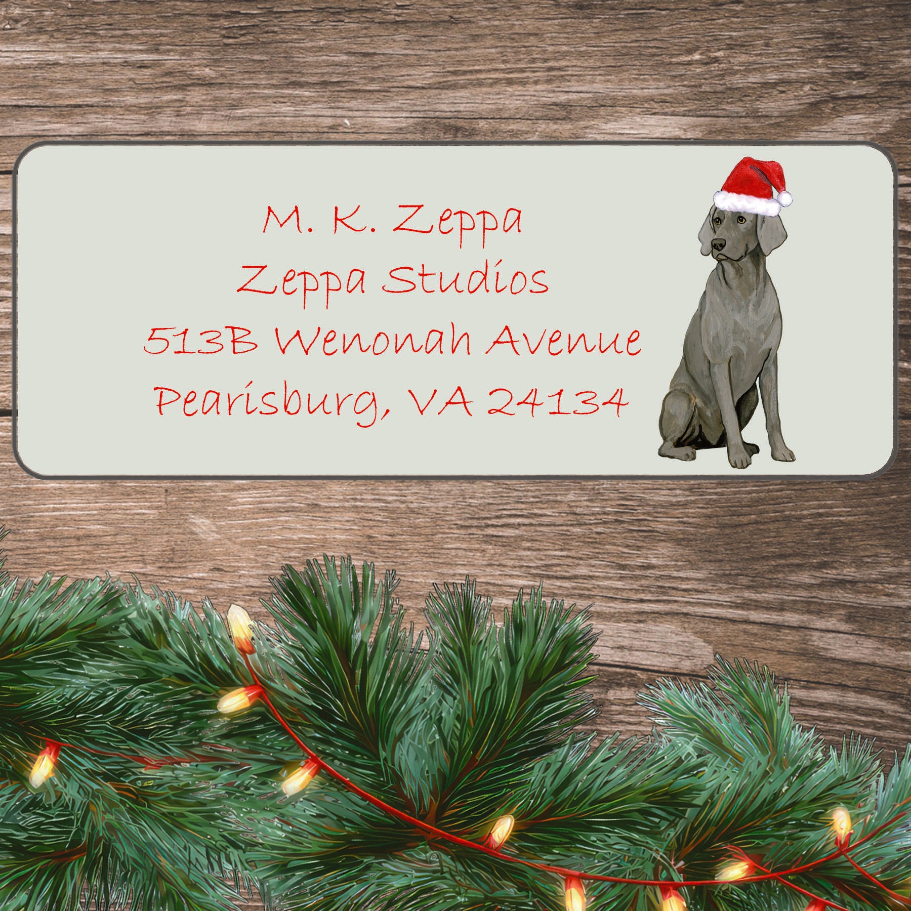 An illustration of a sitting Weimaraner wearing a Santa Hat on an ivory return address label set off by a wood surface with holiday greenery background.