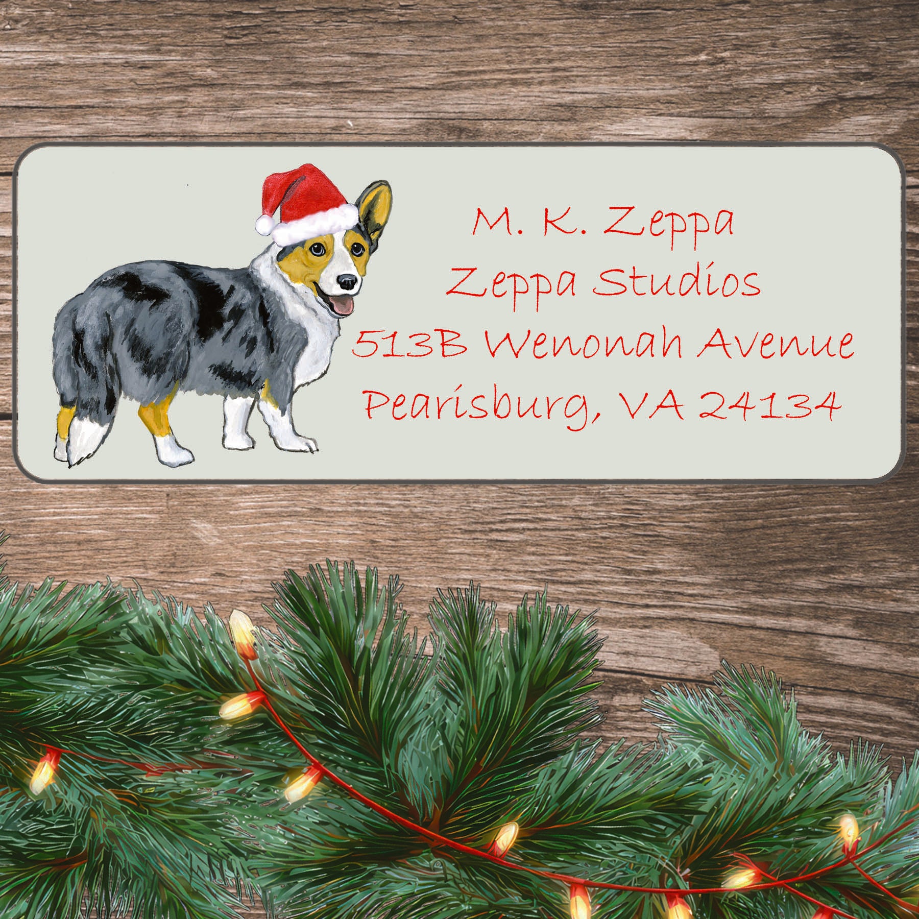 A blue merle Cardigan Welsh Corgi in a Santa Hat illustration on an ivory return address label set off by a wood surface with holiday greenery background.