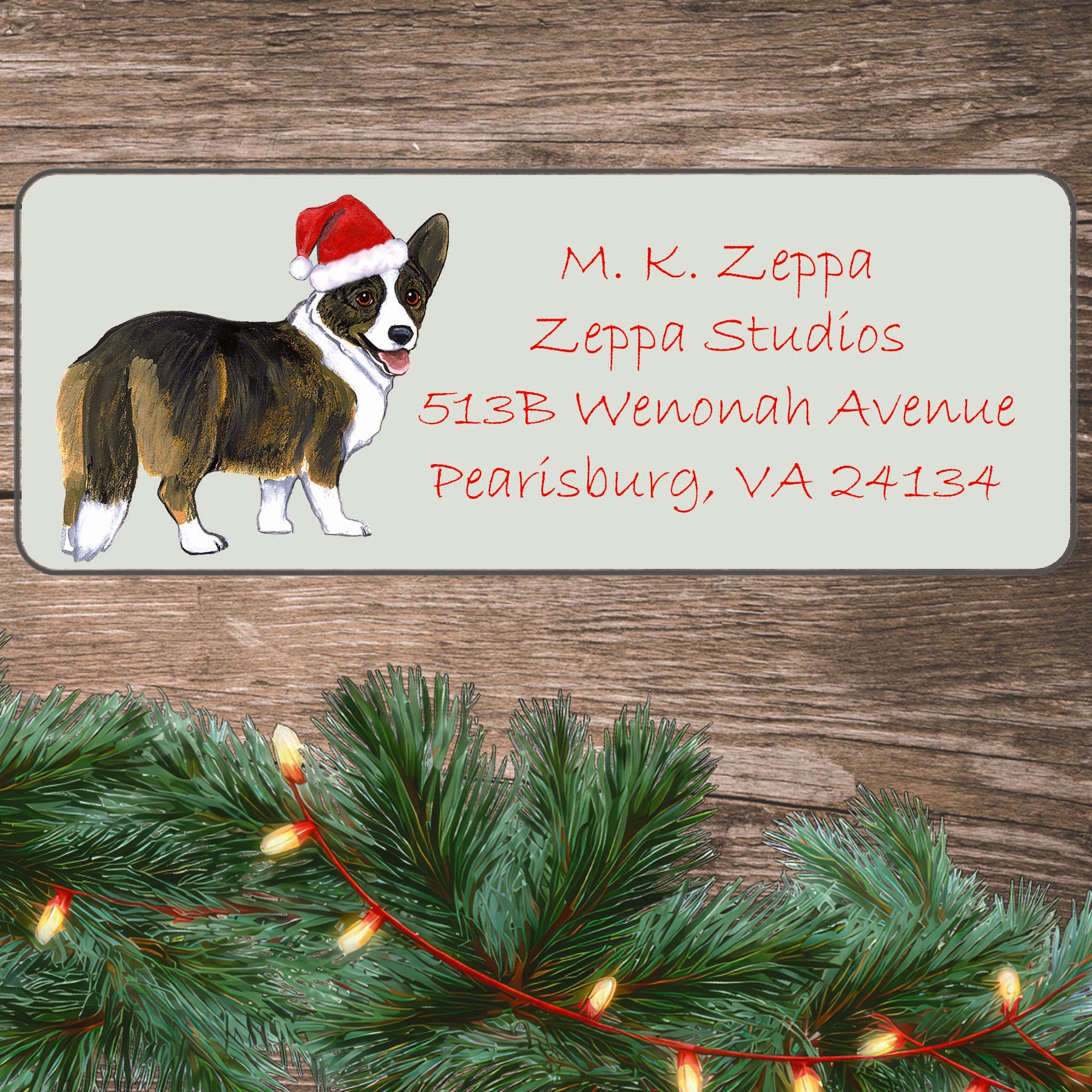 A Cardigan Welsh Corgi in a Santa Hat illustration on an ivory return address label set off by a wood surface with holiday greenery background.
