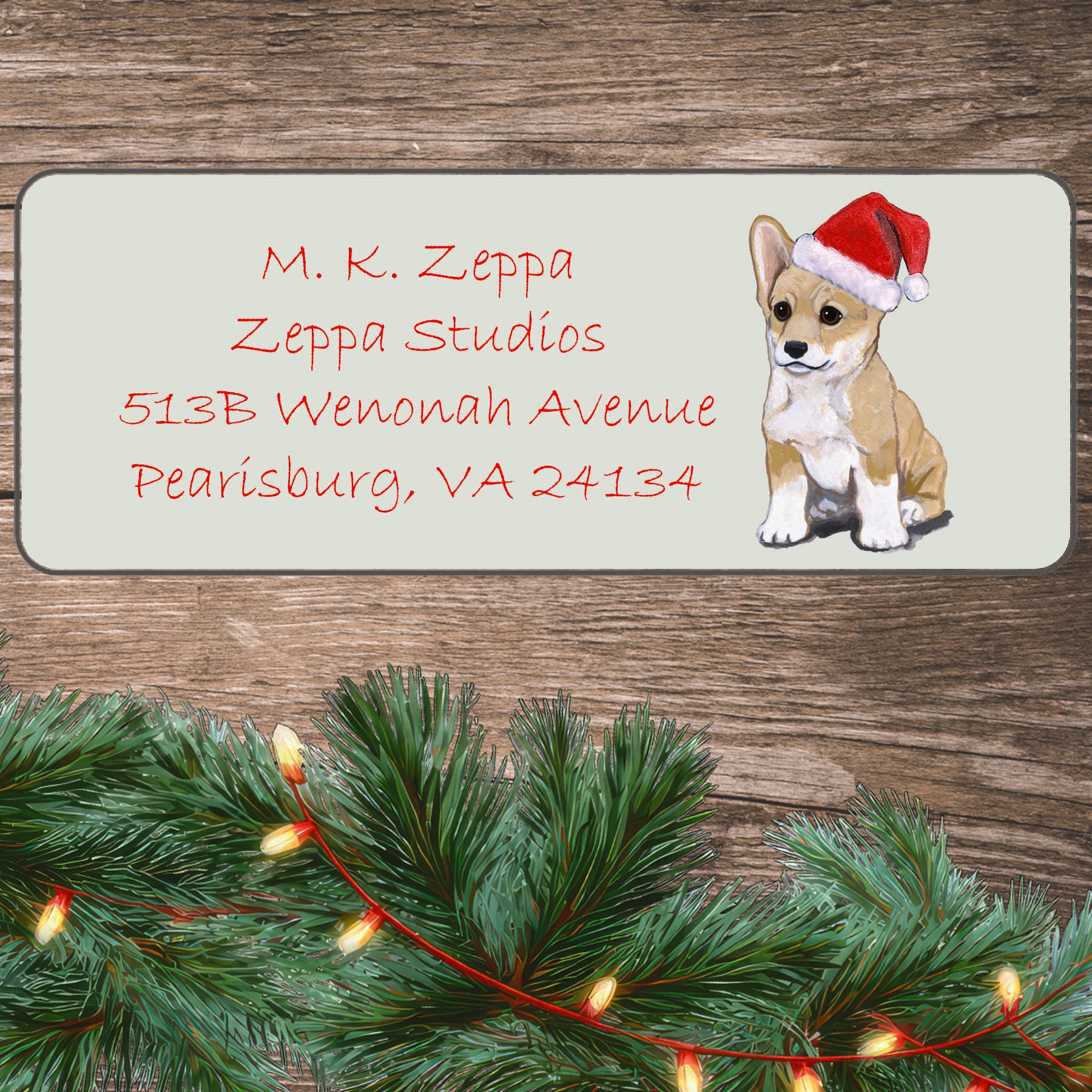 A Pembroke Welsh Corgi puppy in a Santa Hat illustration on an ivory return address label set off by a wood surface with holiday greenery background.