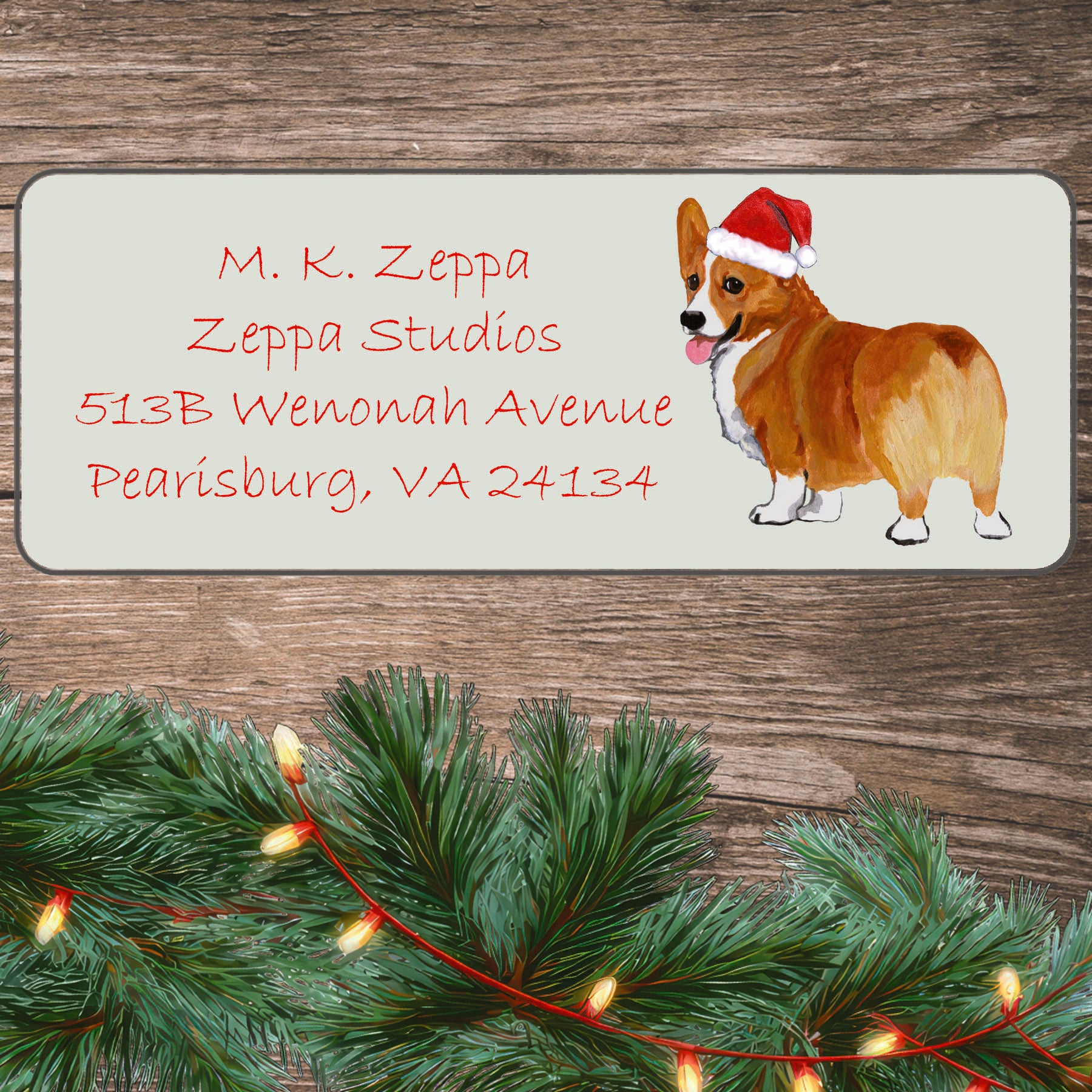 A Pembroke Welsh Corgi in a Santa Hat illustration on an ivory return address label set off by a wood surface with holiday greenery background.