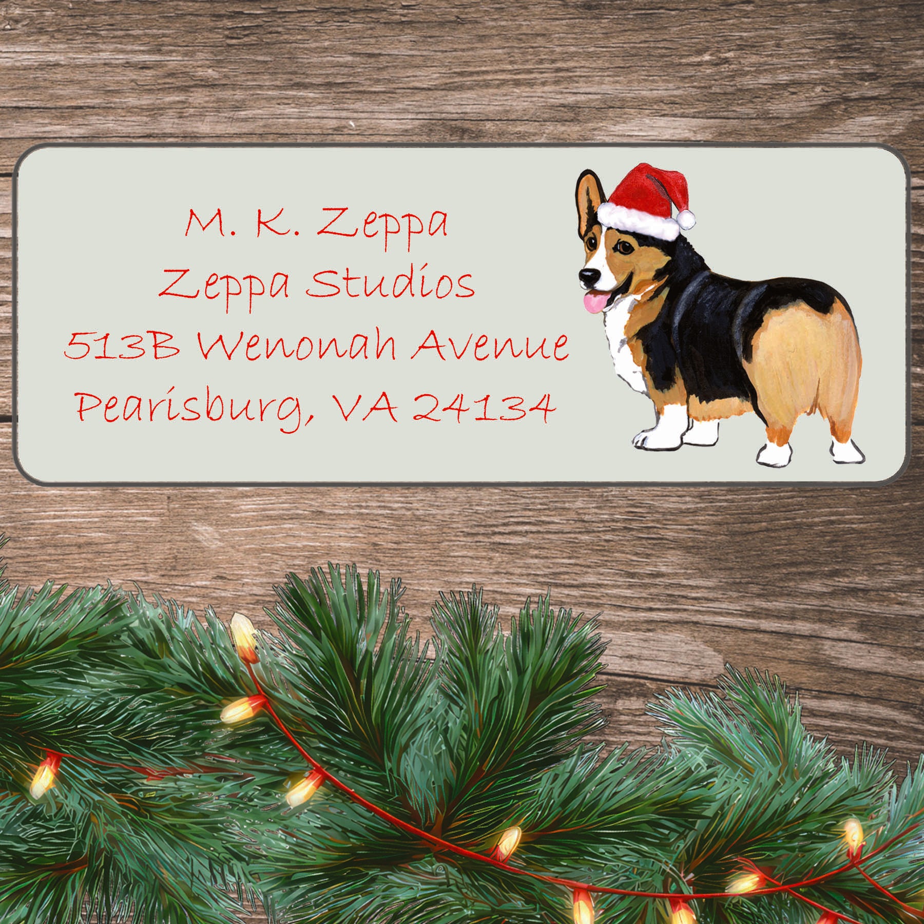 A tri color Pembroke Welsh Corgi in a Santa Hat illustration on an ivory return address label set off by a wood surface with holiday greenery background.
