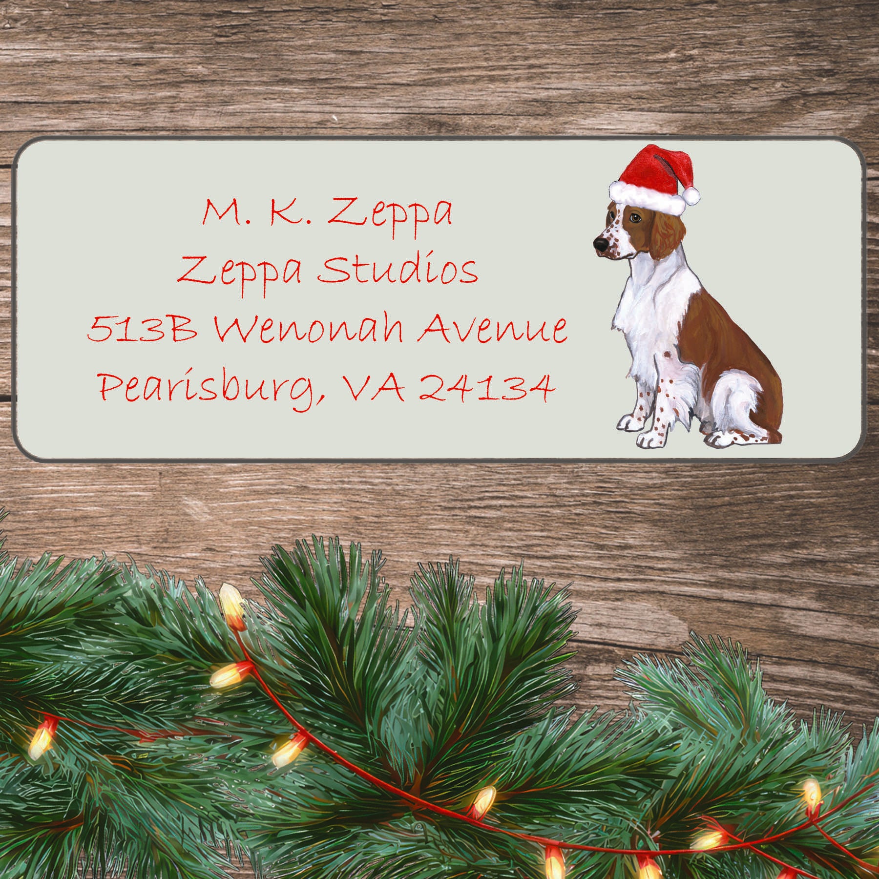 An illustration of a Welsh Springer Spaniel wearing a Santa Hat on an ivory return address label set off by a wood surface with holiday greenery background.