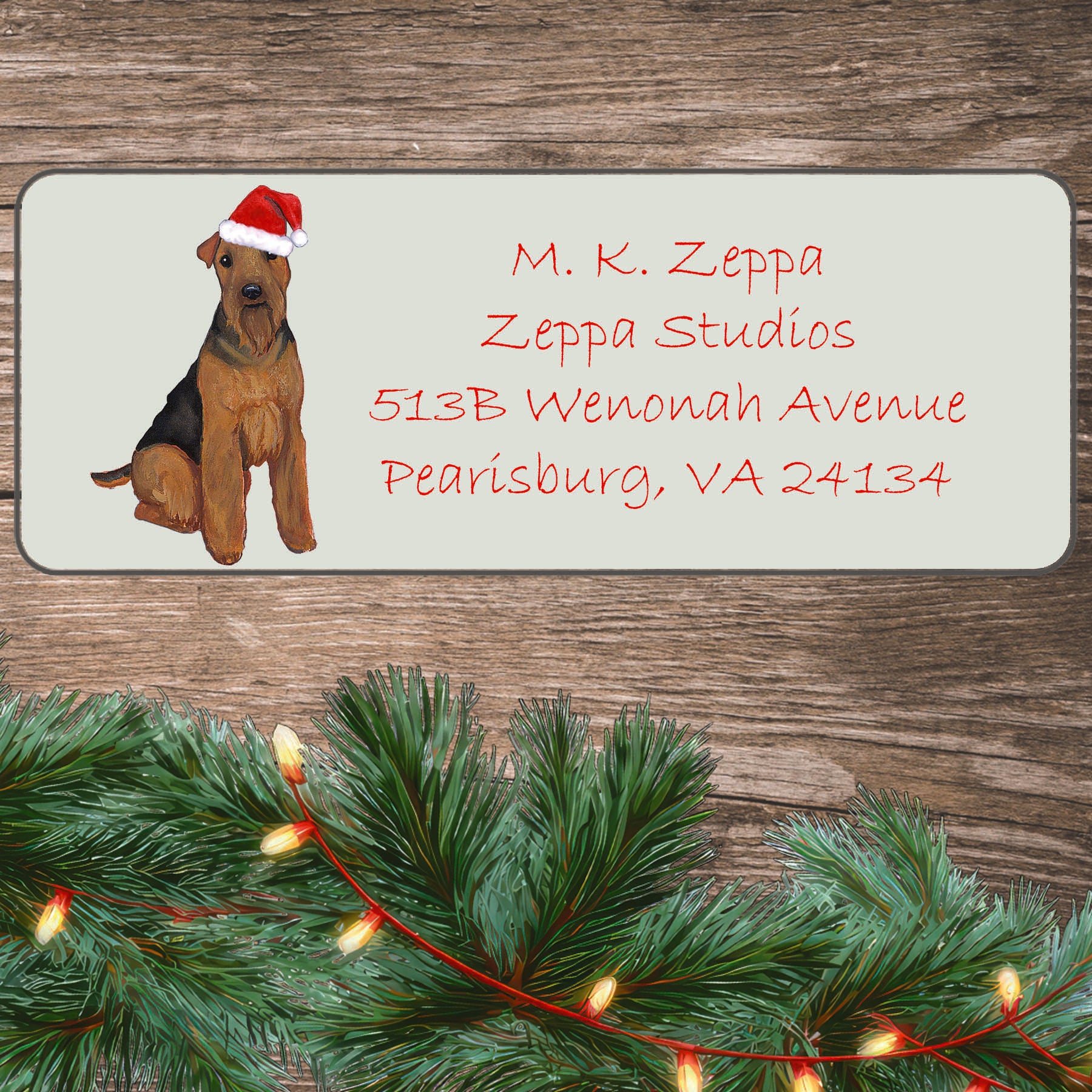 An illustration of a Welsh Terrier wearing a Santa Hat on an ivory return address label set off by a wood surface with holiday greenery background.