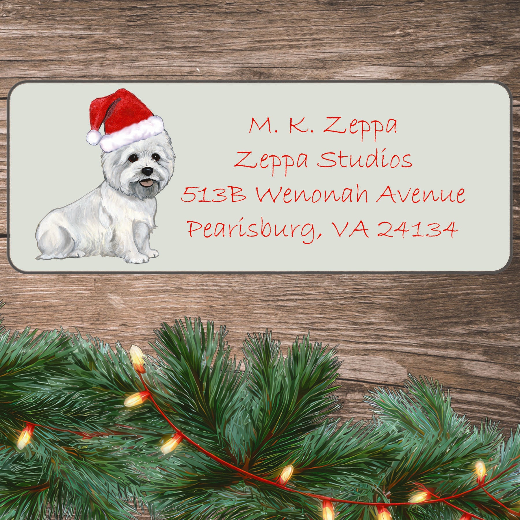An illustration of a sitting West Highland White Terrier wearing a Santa Hat on an ivory return address label set off by a wood surface with holiday greenery background.