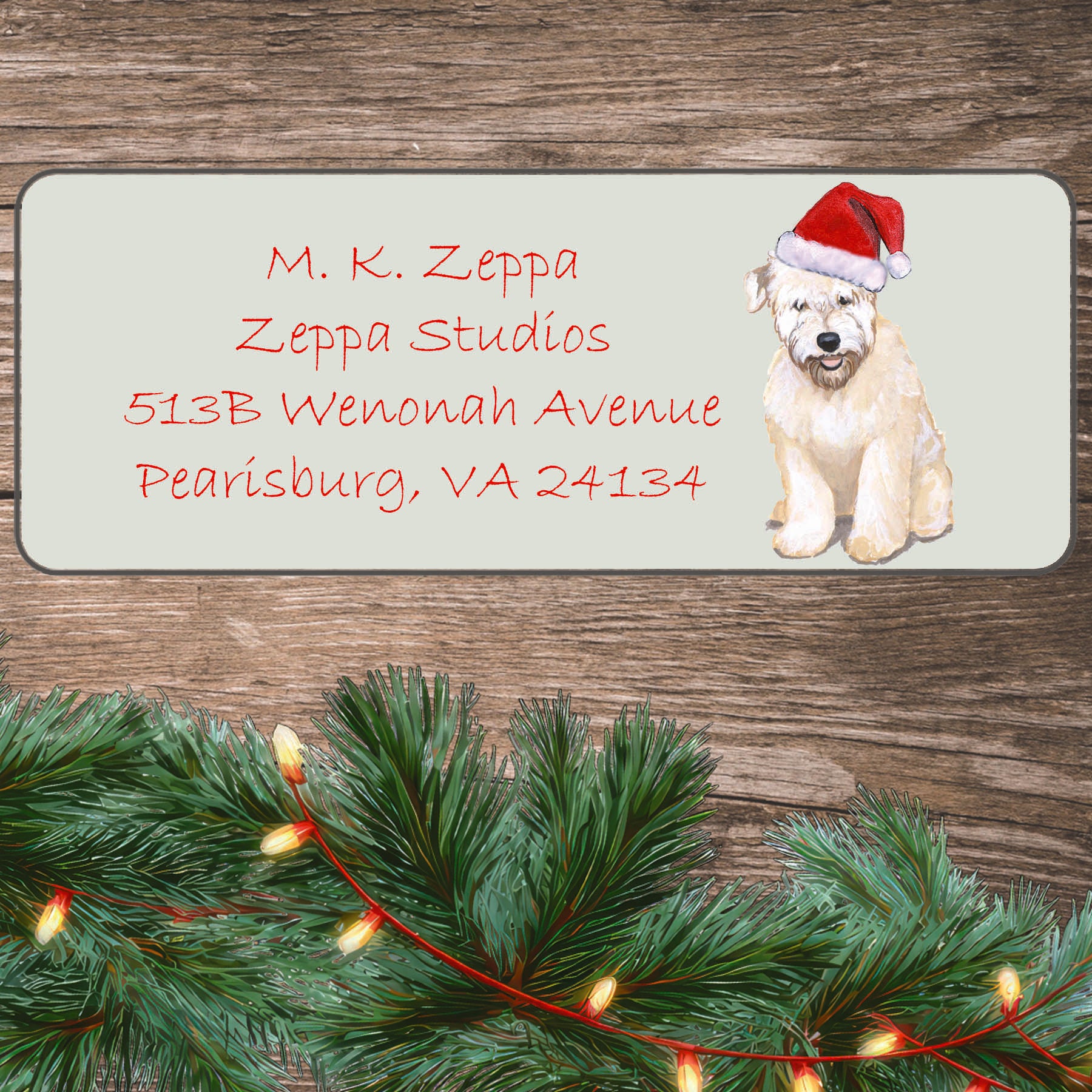 An illustration of a Wheaten Terrier Puppy wearing a Santa Hat on an ivory return address label set off by a wood surface with holiday greenery background.
