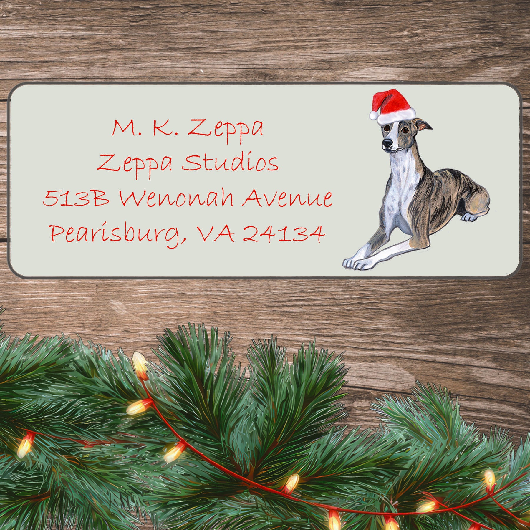 An illustration of a Whippet wearing a Santa Hat on an ivory return address label set off by a wood surface with holiday greenery background.