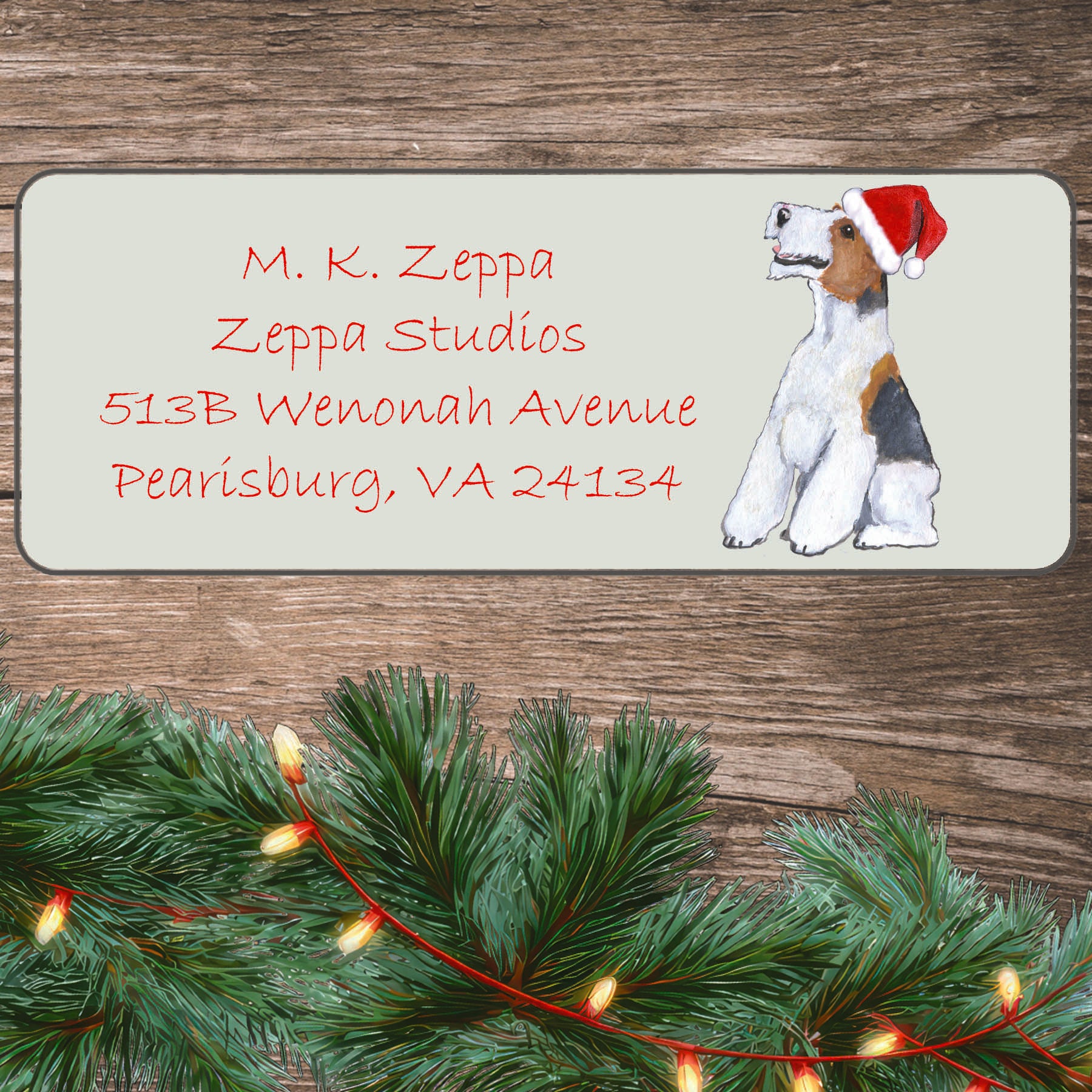 An illustration of a Wire Fox Terrier wearing a Santa Hat on an ivory return address label set off by a wood surface with holiday greenery background.