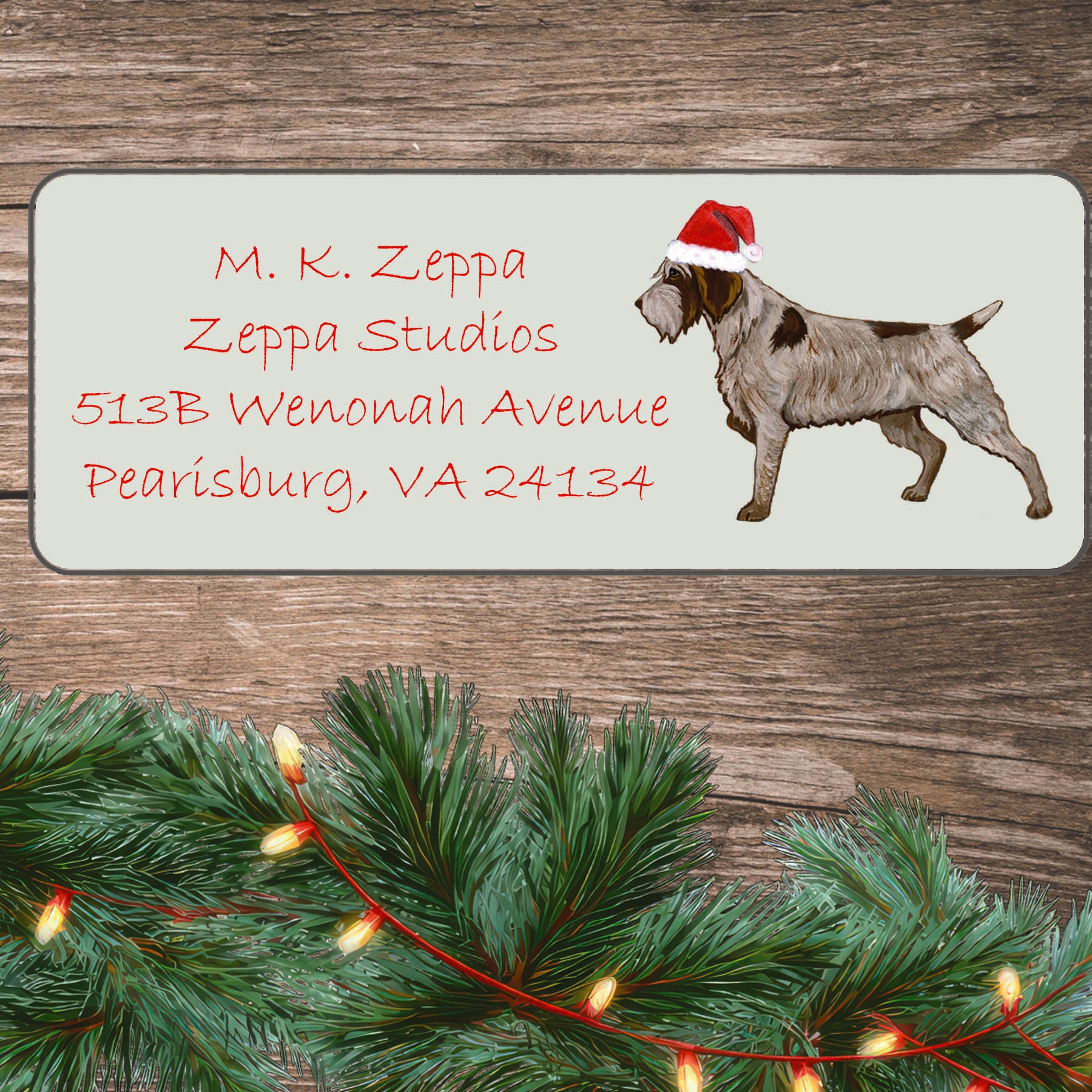 An illustration of a Wirehaired Pointing Griffon wearing a Santa Hat on an ivory return address label set off by a wood surface with holiday greenery background.