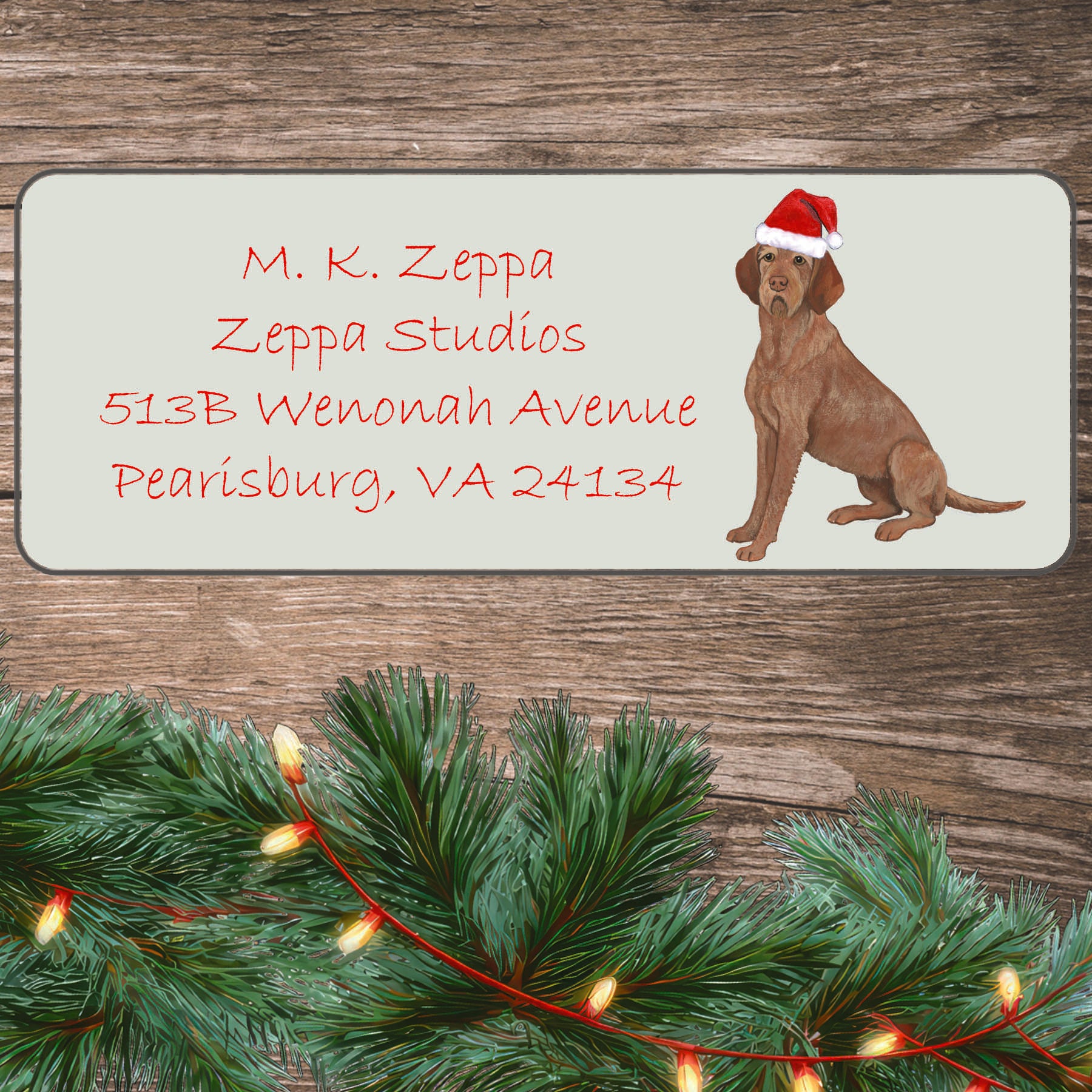 Wirehaired Vizsla in a Santa Hat on an ivory return address label set off by a wood surface with holiday greenery background.