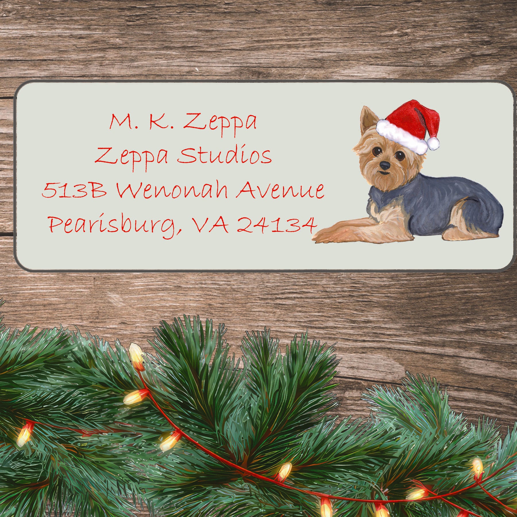 An illustration of a Yorkie with a short haircut wearing a Santa Hat on an ivory return address label set off by a wood surface with holiday greenery background.