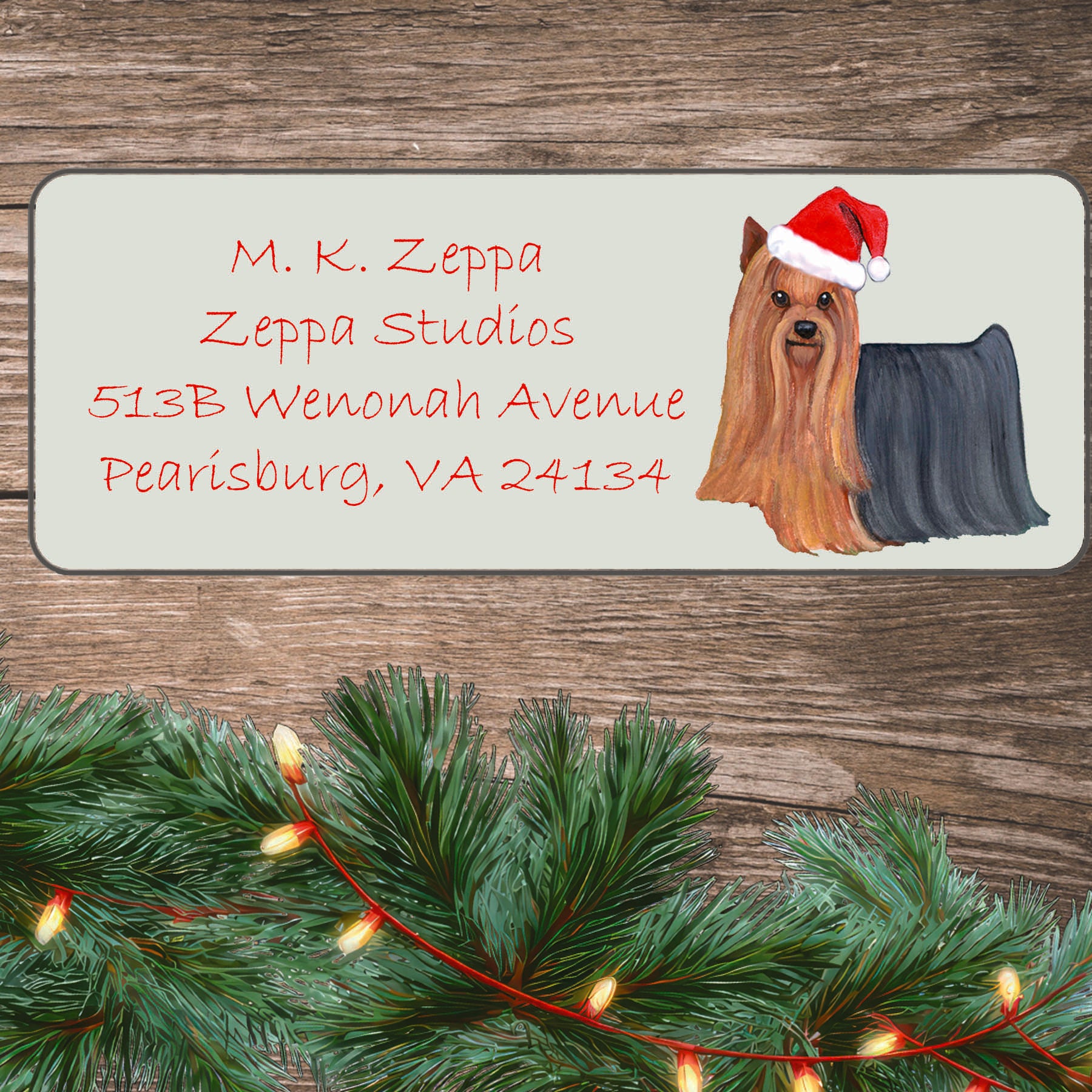 An illustration of a Yorkshire Terrier wearing a Santa Hat on an ivory return address label set off by a wood surface with holiday greenery background.