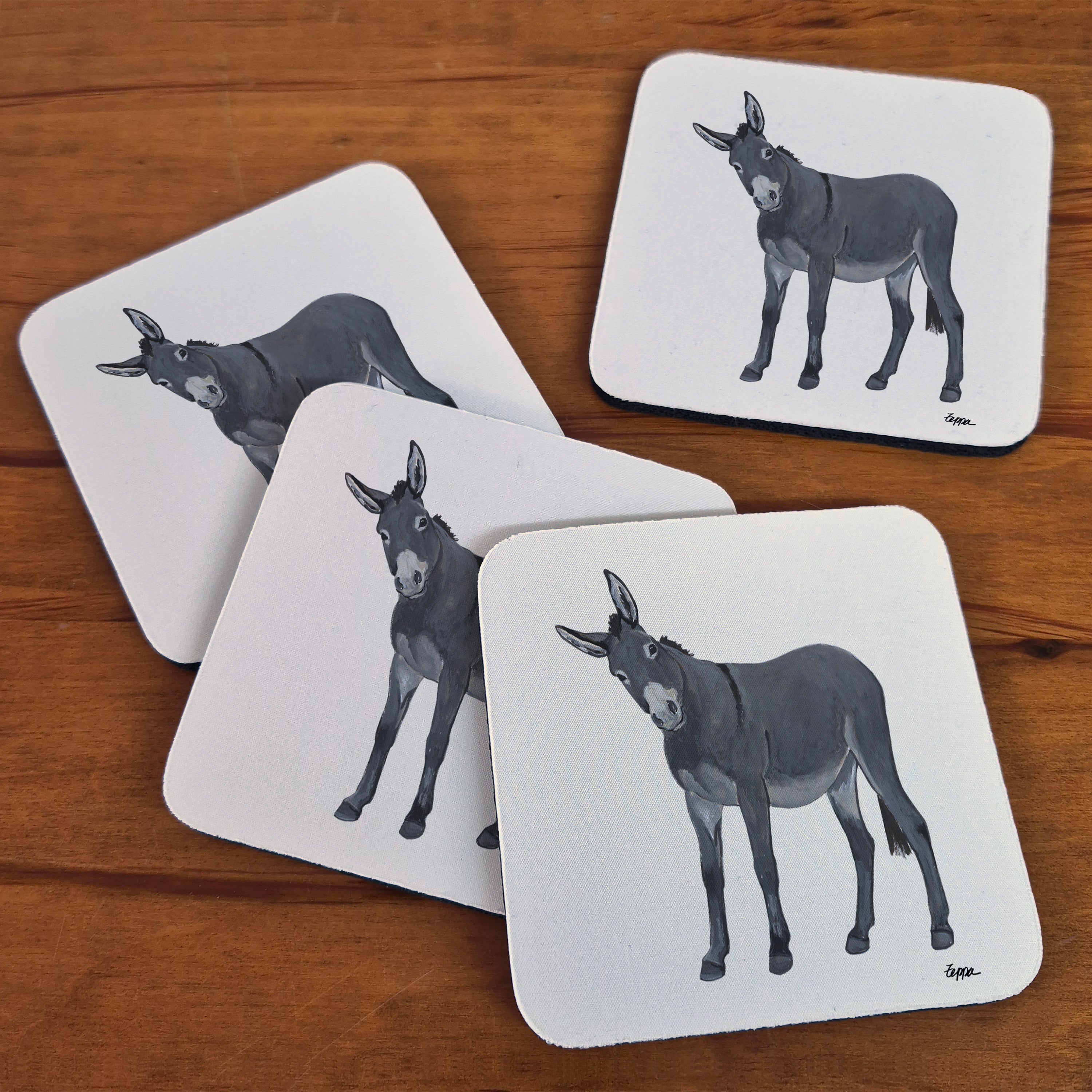 Four square beige coasters with rounded corners, each printed with an illustration of a donkey, arranged on a wooden tabletop.