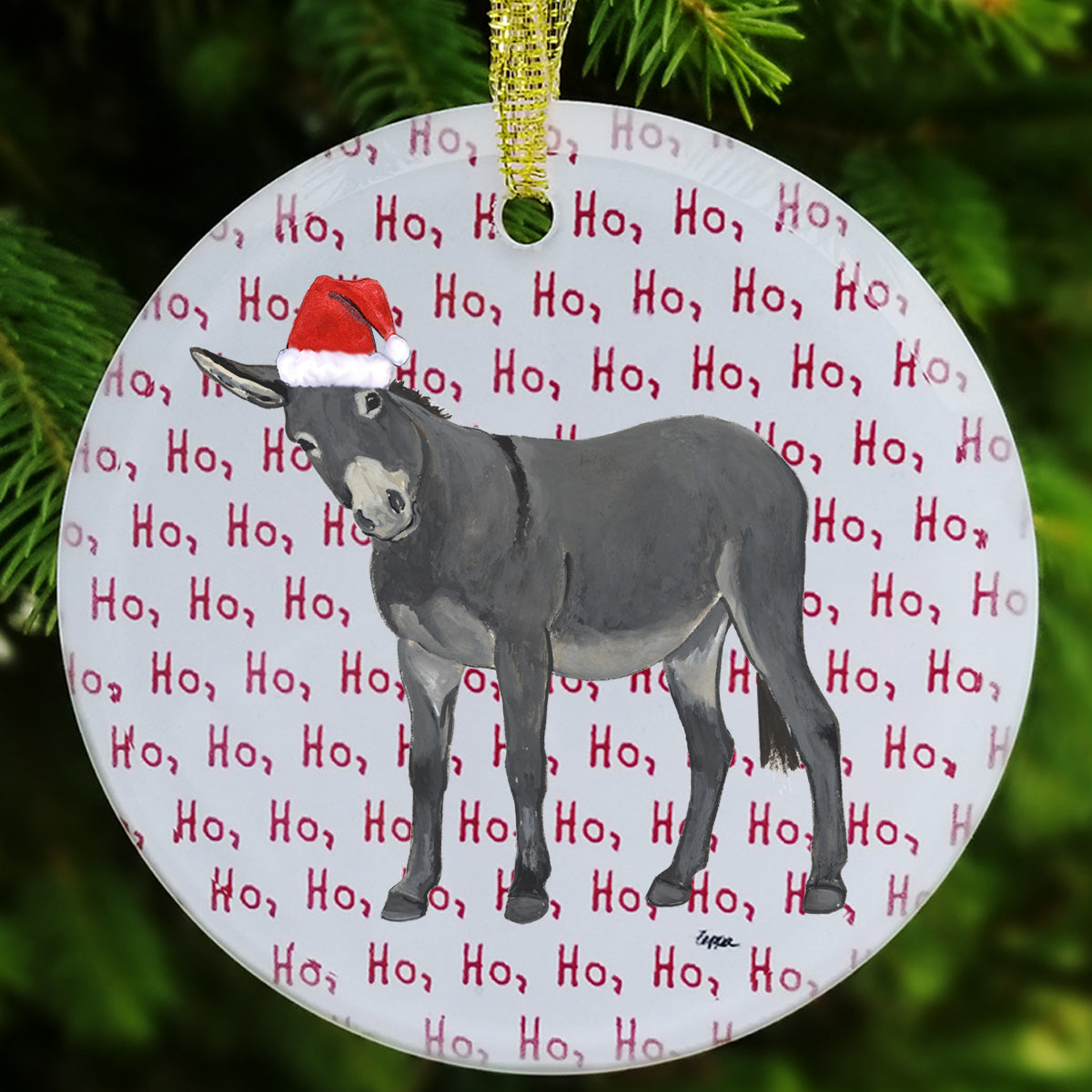 A round glass Christmas ornament featuring a gray donkey wearing a red Santa hat, set against a white background with repeating red 'Ho, Ho, Ho' text. The ornament hangs from a gold string against a blurred green pine background.