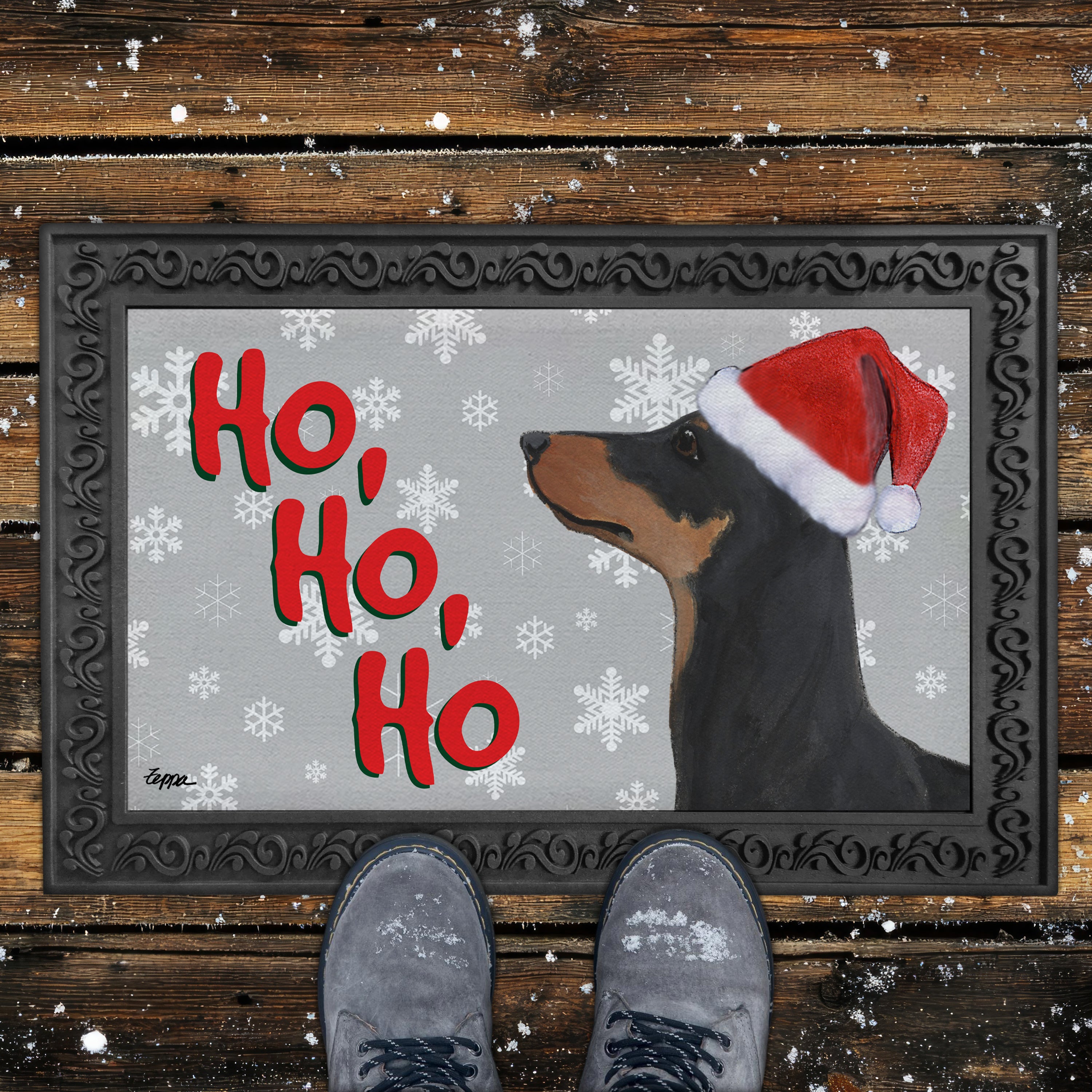 German Pinscher in a Santa hat on a Doormat with a grey background with Ho Ho Ho text and white snowflakes.