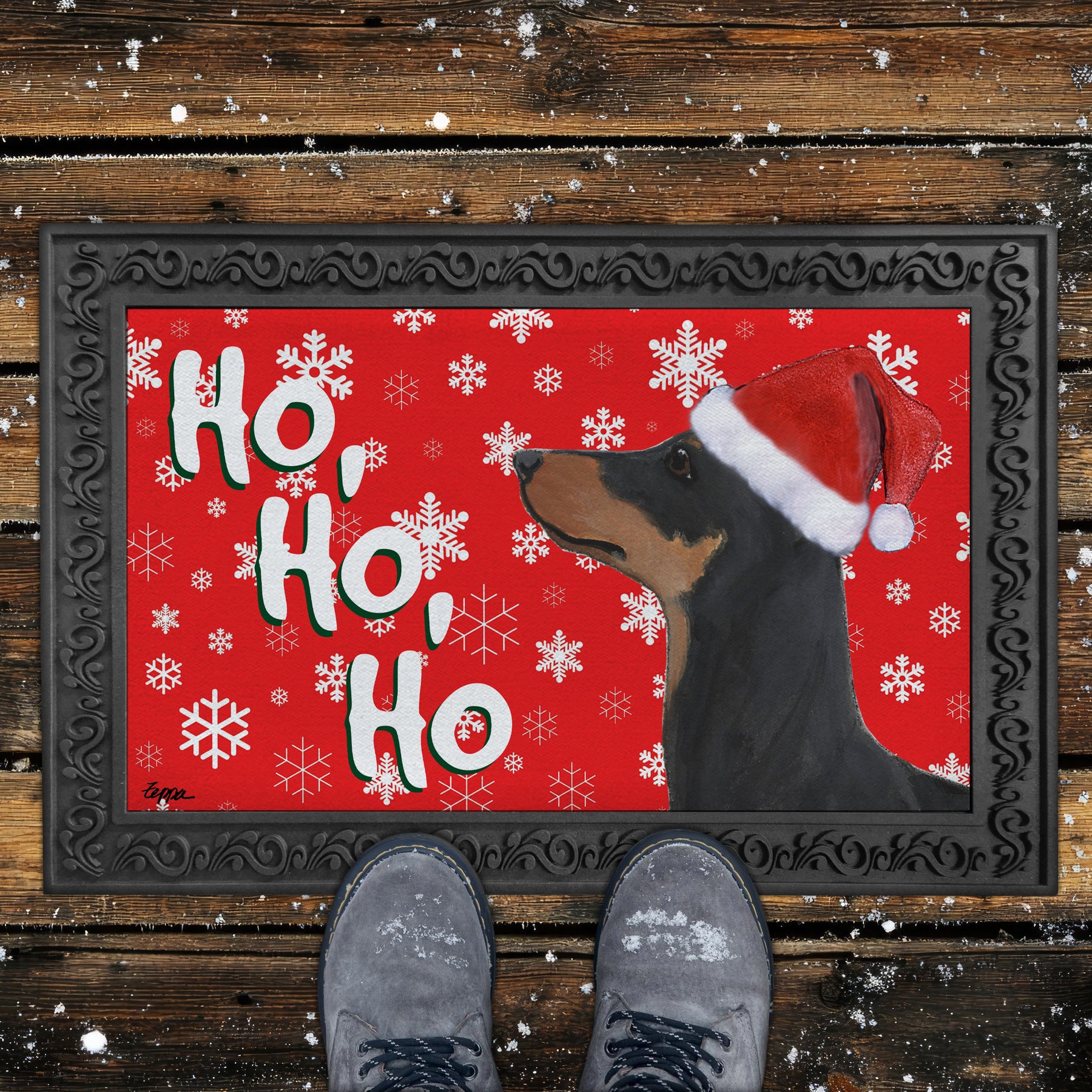 German Pinscher in a Santa hat on a Doormat with a red background with Ho Ho Ho text and white snowflakes.
